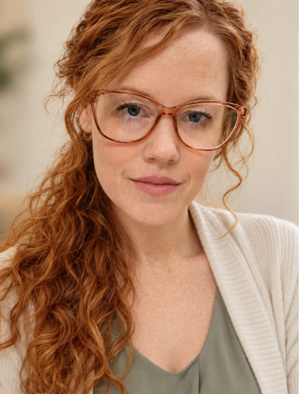 Headshot of Sara Murphy, LMSW, therapist at Upstate Counseling, with long curly red hair and freckles, wearing glasses, a sage green top, and a cream-colored cardigan against a softly blurred neutral background.
