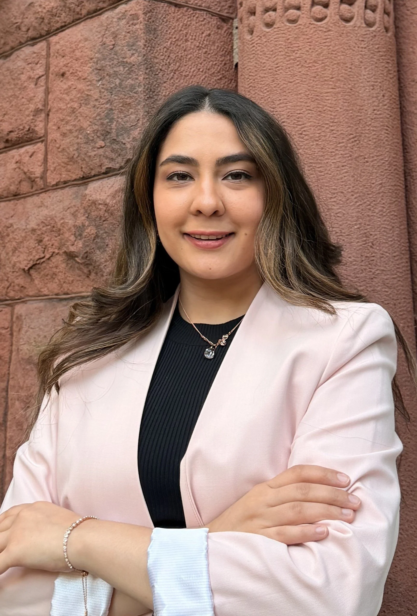 Headshot of Mariyam Moqbil, MHC-LP, therapist at Upstate Counseling, smiling with long dark wavy hair, wearing a light pink blazer over a black top with her arms crossed, photographed outdoors against a textured stone wall.