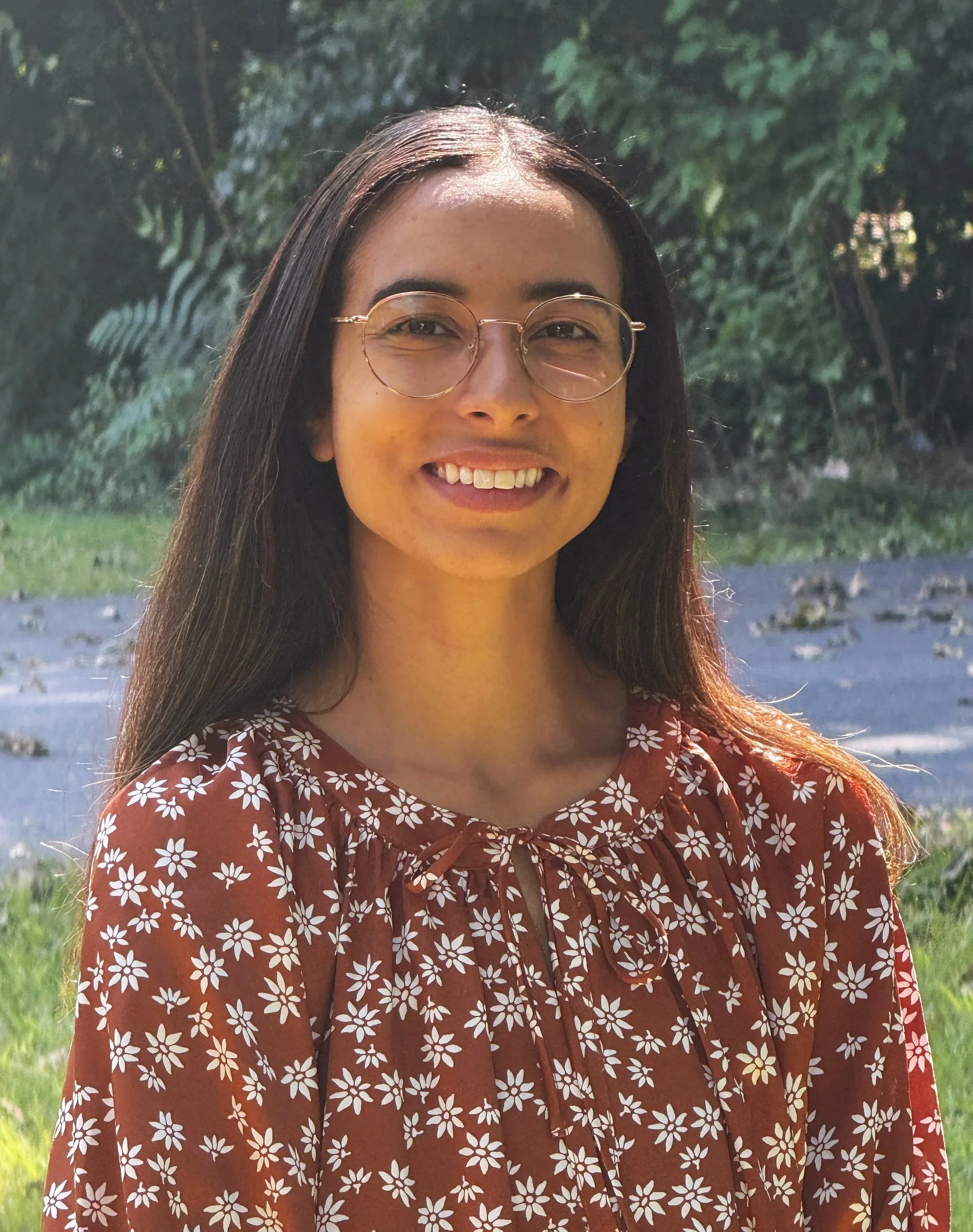 Headshot of Audrey Blake, LMSW, therapist at Upstate Counseling, smiling with long straight dark hair and glasses, wearing a rust-colored floral top, photographed outdoors with a leafy green background.