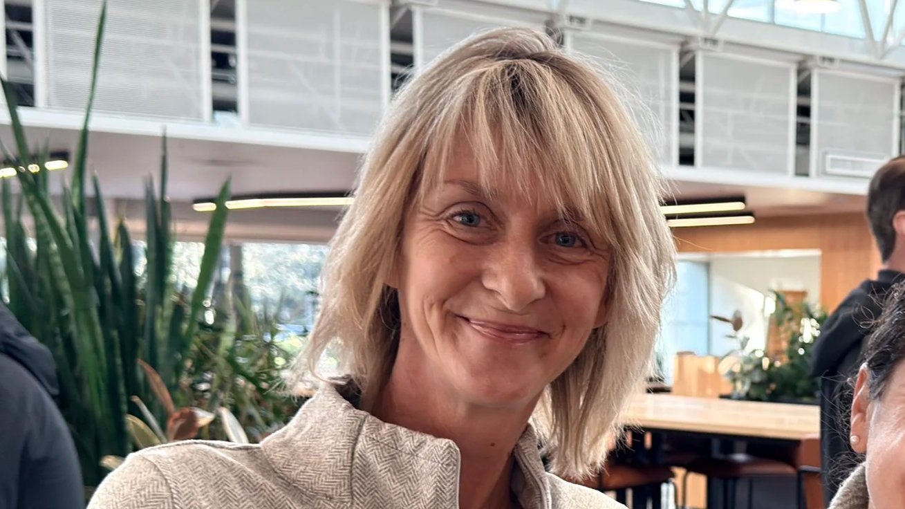 A woman with shoulder-length blonde hair smiling at the camera in a modern indoor setting, with plants and other people in the background.