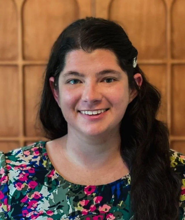 A young woman with long dark hair, smiling, wearing a colorful patterned blouse, standing in front of a wooden panel background.