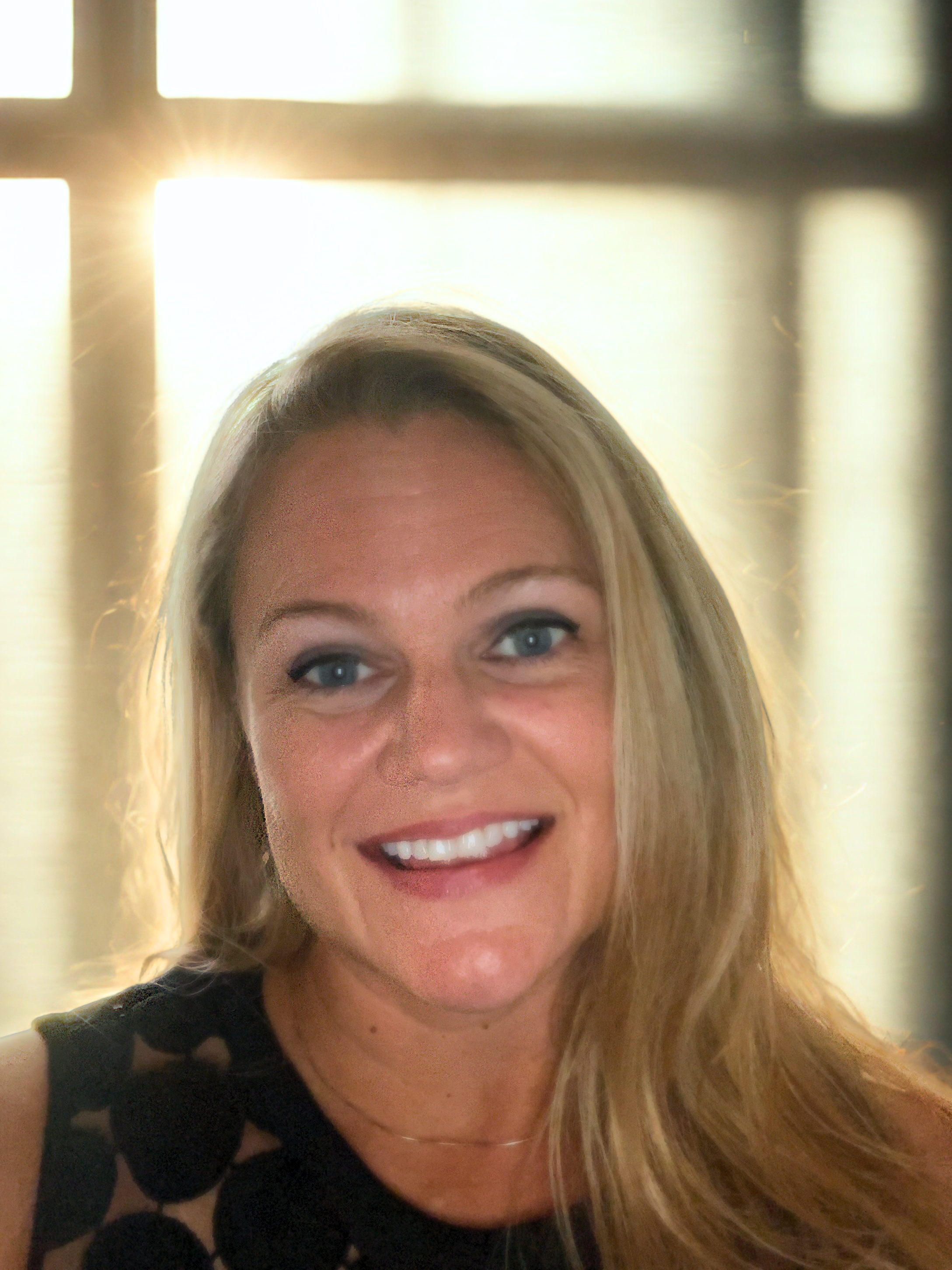 Close-up of a smiling woman with blonde hair and blue eyes, wearing a black top with cut-out detailing, in front of a window with sunlight streaming through.