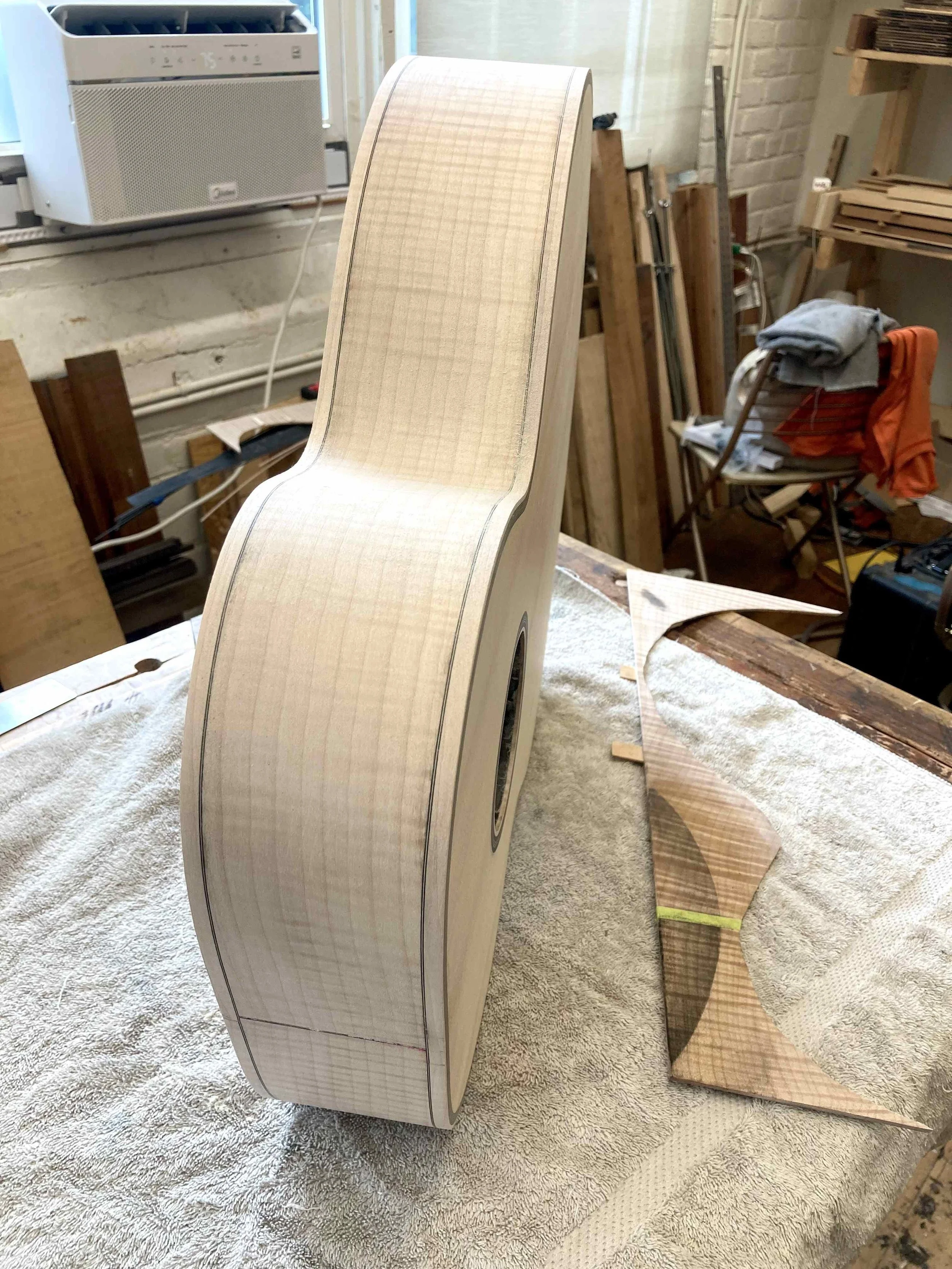 a figured maple guitar rests on the bench of guitar maker Aaron Foster next to a small finish experiment.  the maple has been finished with shellac to create a sealed border and then linseed oil with lamp black in two ways to try and create a burst