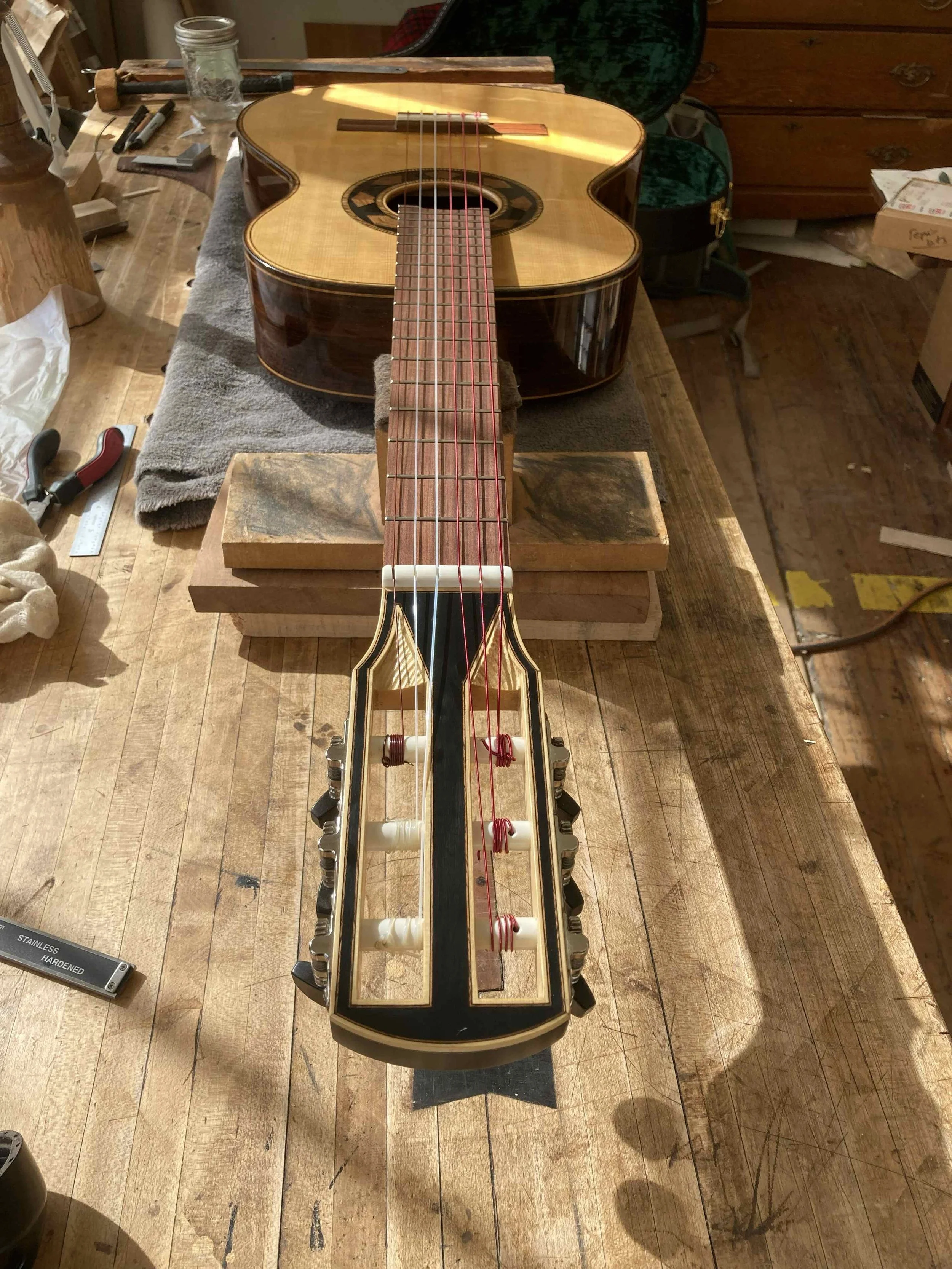 A classical guitar headstock in foreground featuring sculptural scalloped string cutouts transitioning into square tuning machine slots that appear bound.  The lampblack and oil finish it bordered by purfling and contrasts with bright yellow cedar