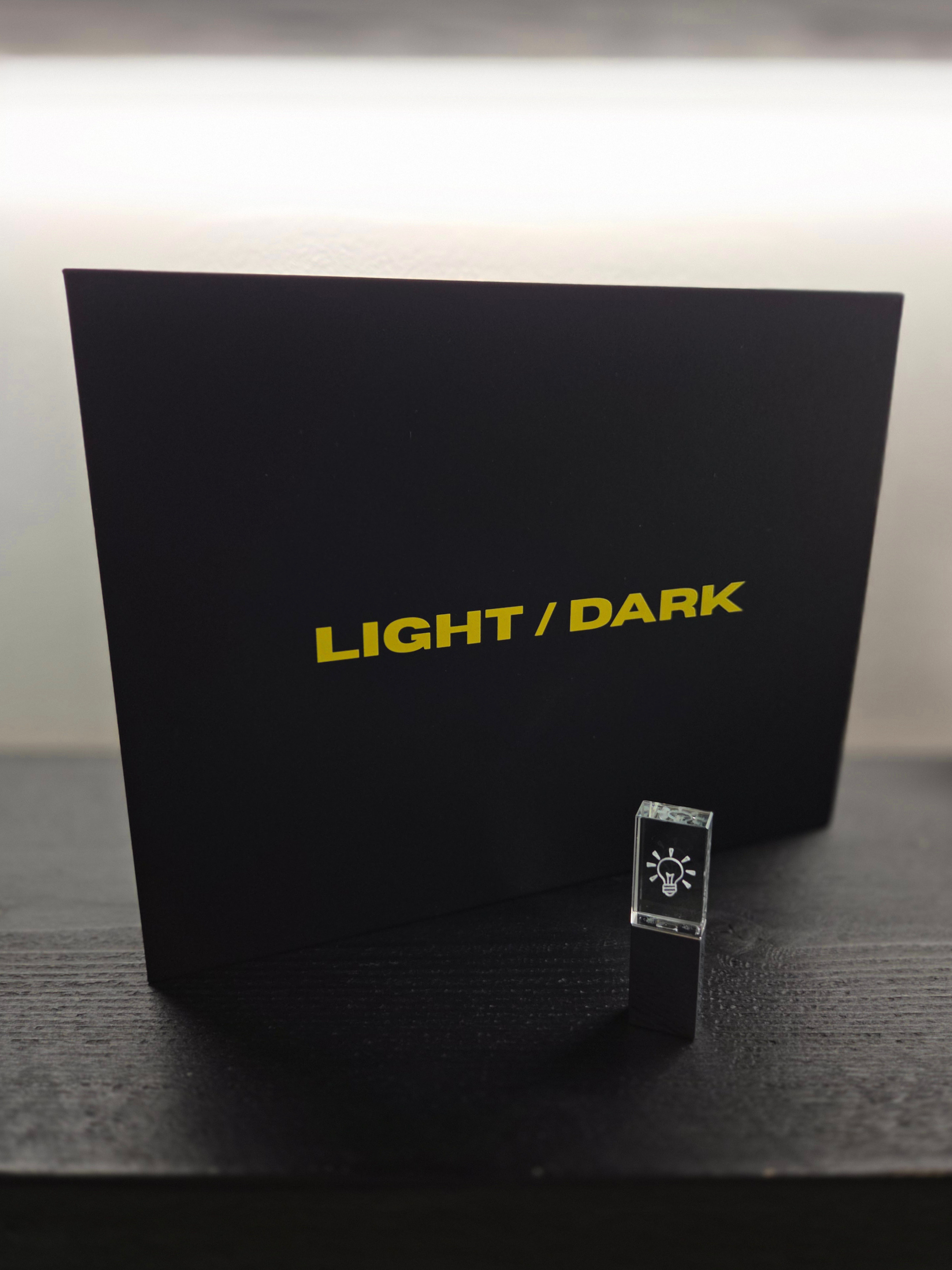 A black box with yellow text 'LIGHT / DARK' and a small transparent cube with a lightbulb icon in front of it on a black wooden surface.