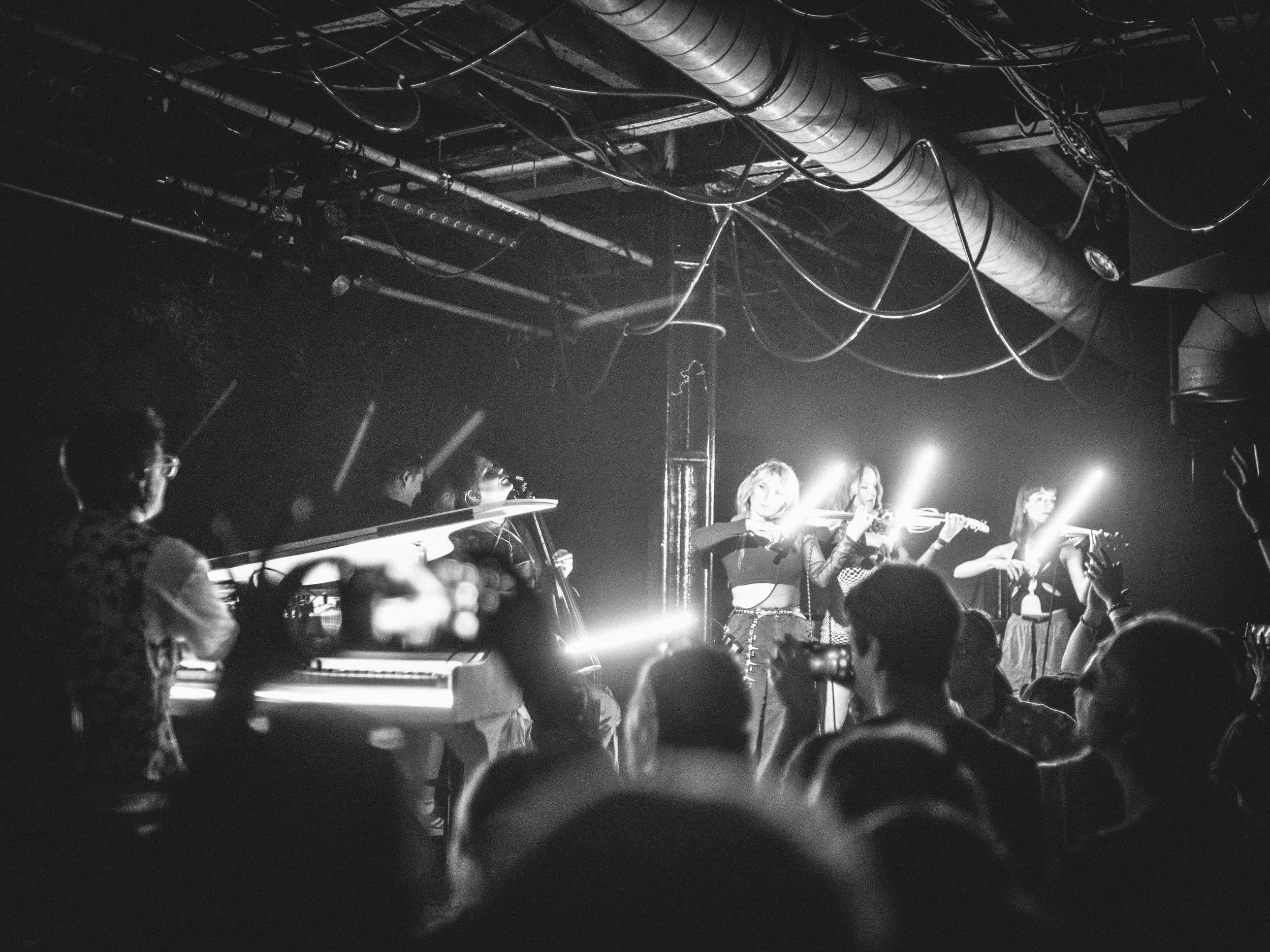 Black and white photo of a crowd at a concert or event, with people in the foreground and a stage with bright lights in the background.