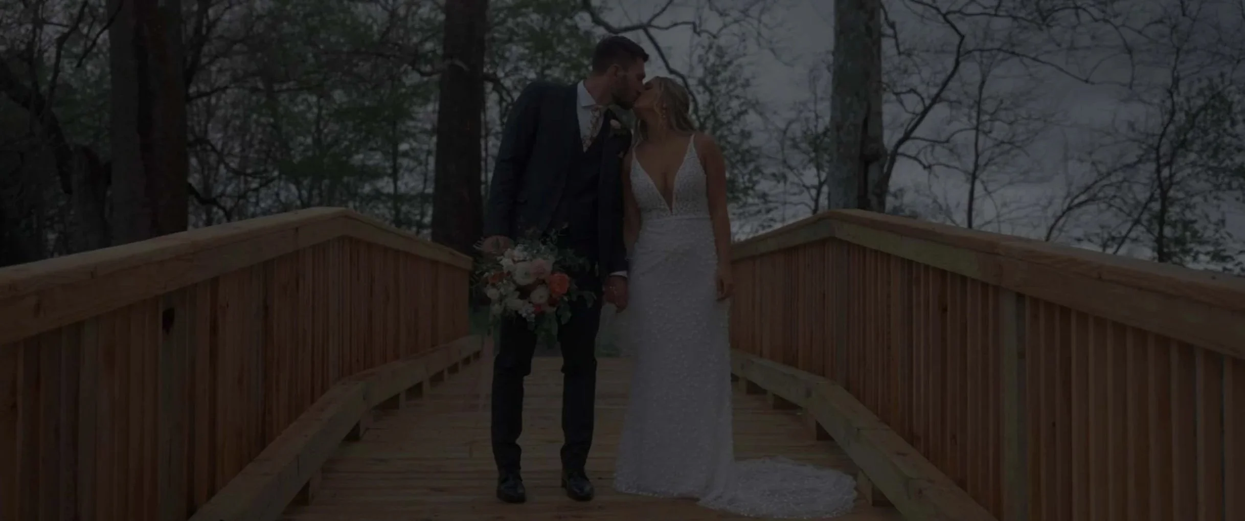 A newlywed couple shares a kiss on a wooden bridge outdoors surrounded by trees, with the bride wearing a white beaded wedding gown and the groom in a dark suit holding a bouquet of flowers.
