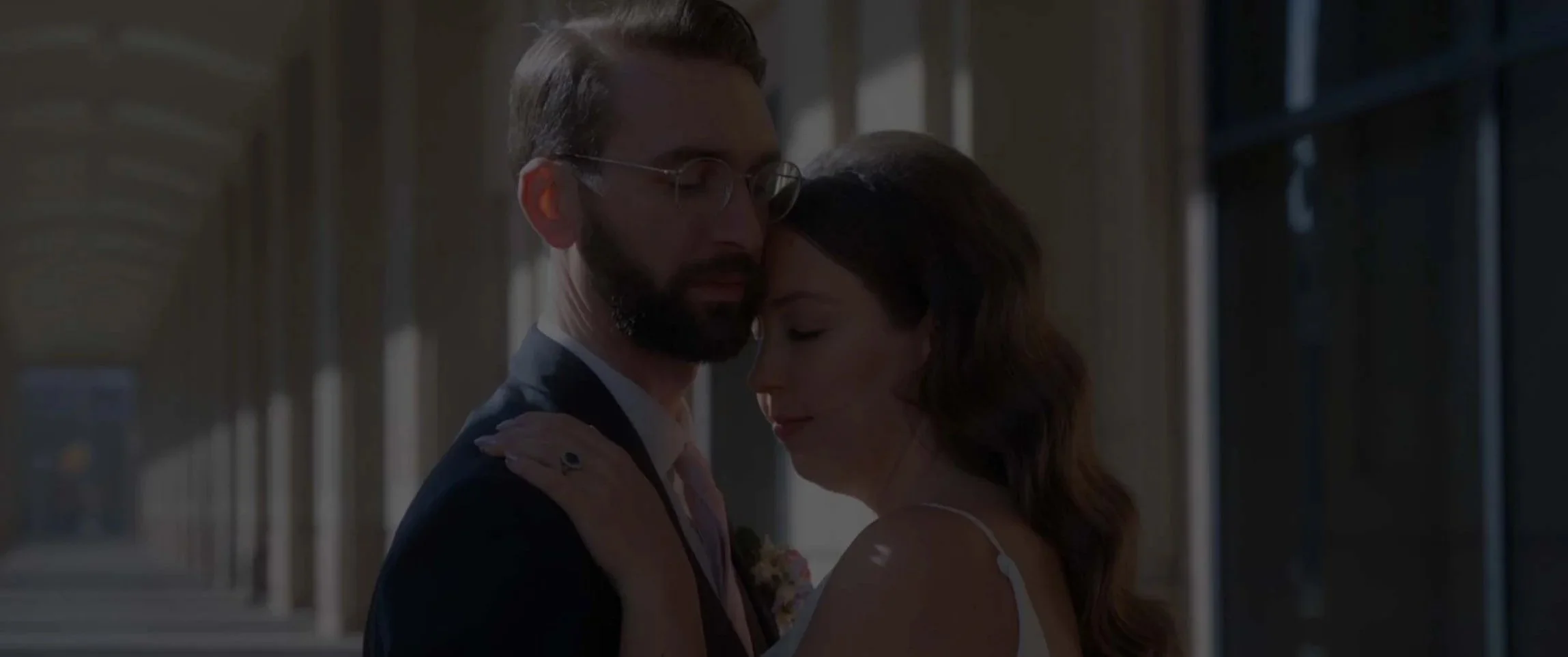 A couple sharing an intimate moment, with the man in a suit and the woman in a wedding dress, in a softly lit indoor setting.