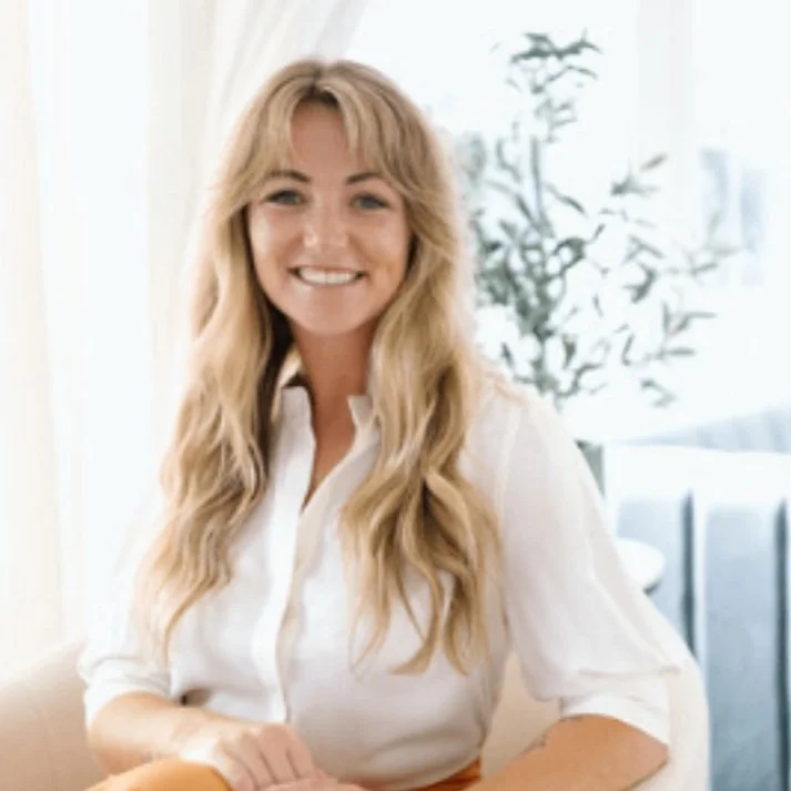 A smiling woman with long, wavy blonde hair wearing a white blouse, sitting in a bright room with a plant and a sofa in the background.