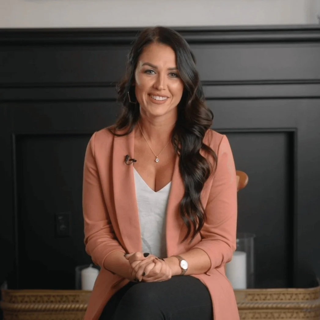 A woman with long dark wavy hair, wearing a light pink blazer over a white top, sitting in front of a black fireplace, smiling and looking at the camera.  Wedding planner