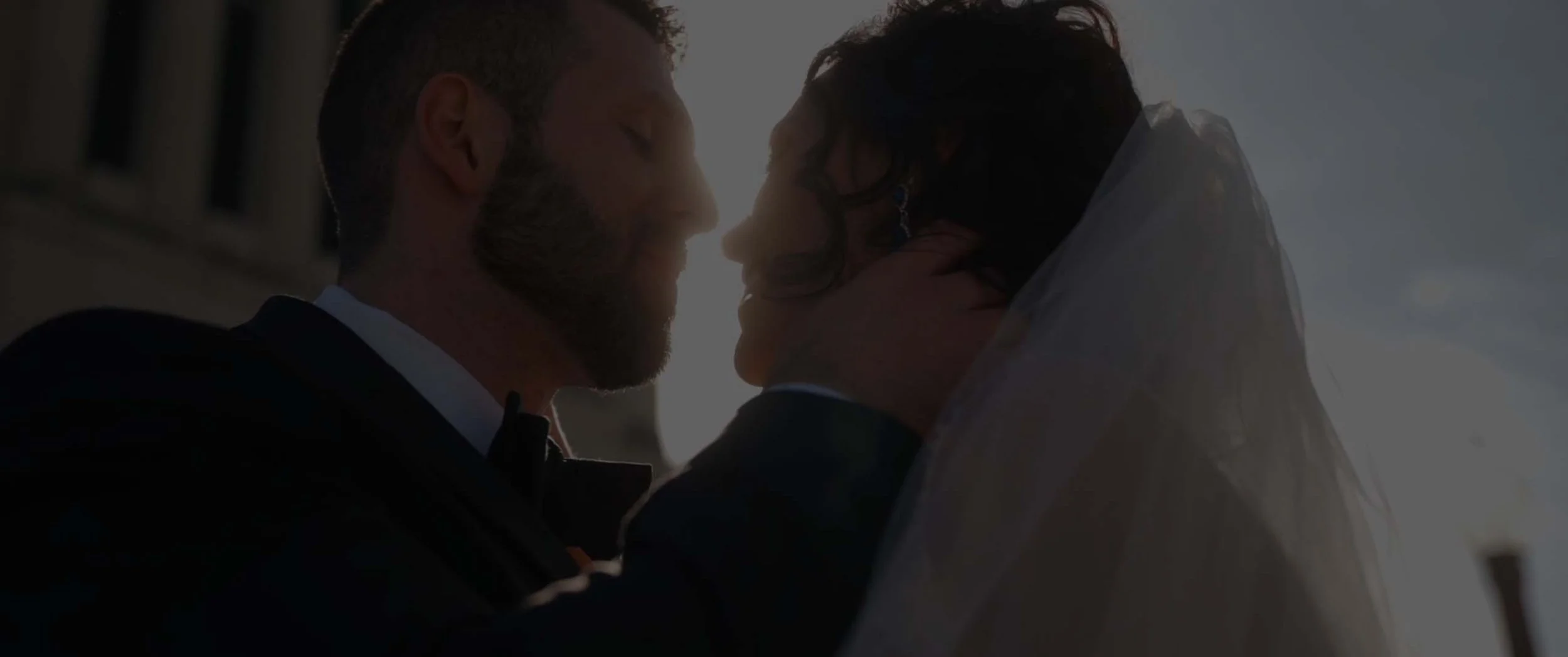 A close-up of a couple at sunset, about to kiss, with their faces very close together. The man has a beard and is wearing a tuxedo, and the woman has dark hair, earrings, and is dressed in formal attire.