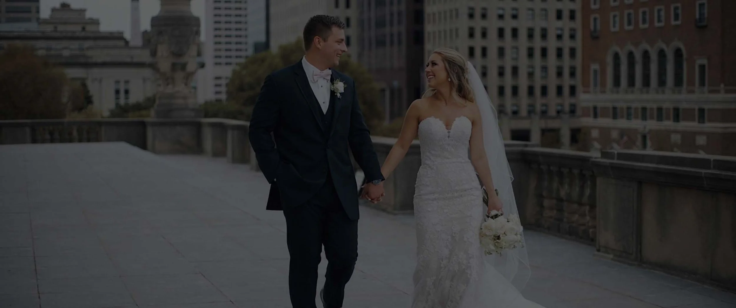 A bride and groom walking hand-in-hand outdoors in a city, smiling at each other. The groom is wearing a black tuxedo with a pink bow tie, and the bride is wearing a white lace wedding gown, holding a bouquet of white roses. Indianapolis Wedding