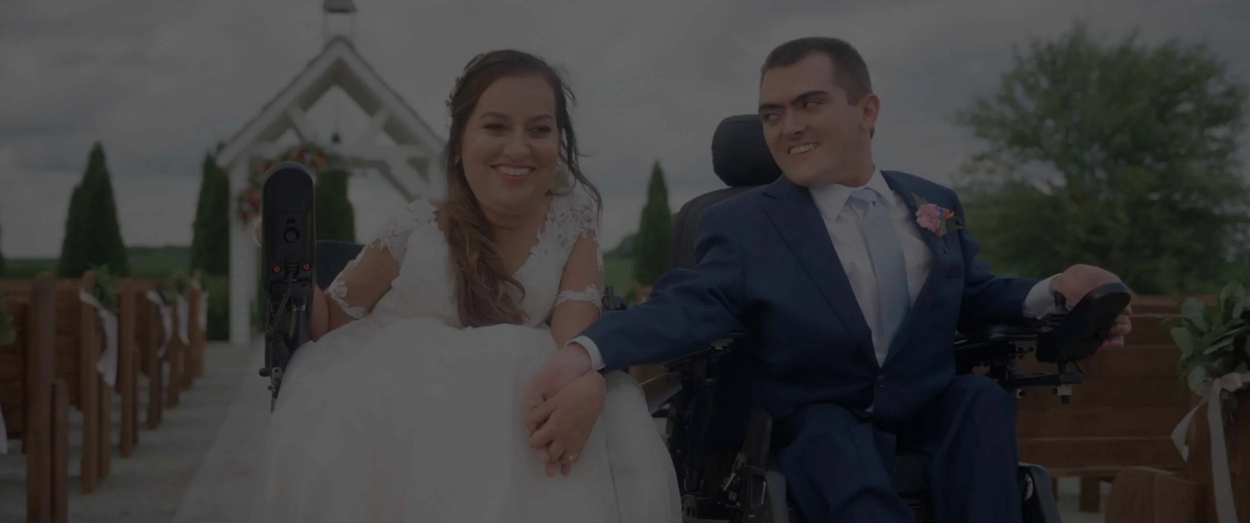 A bride and groom, one in a wedding dress and the other in a suit, holding hands and smiling at each other outdoors.
