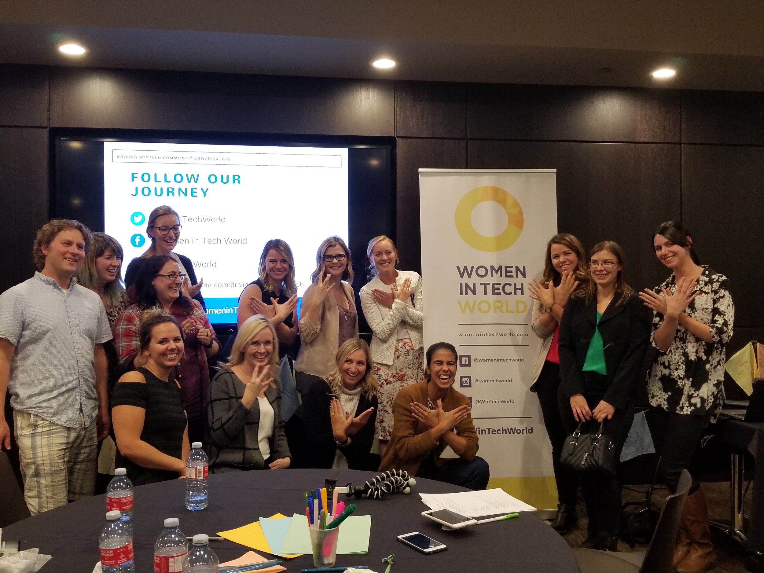  Image is of the Community Conversation in Saskatoon. There is a group of people gathered in front of the WiTWorld sign in a conference room, posing for a picture.  