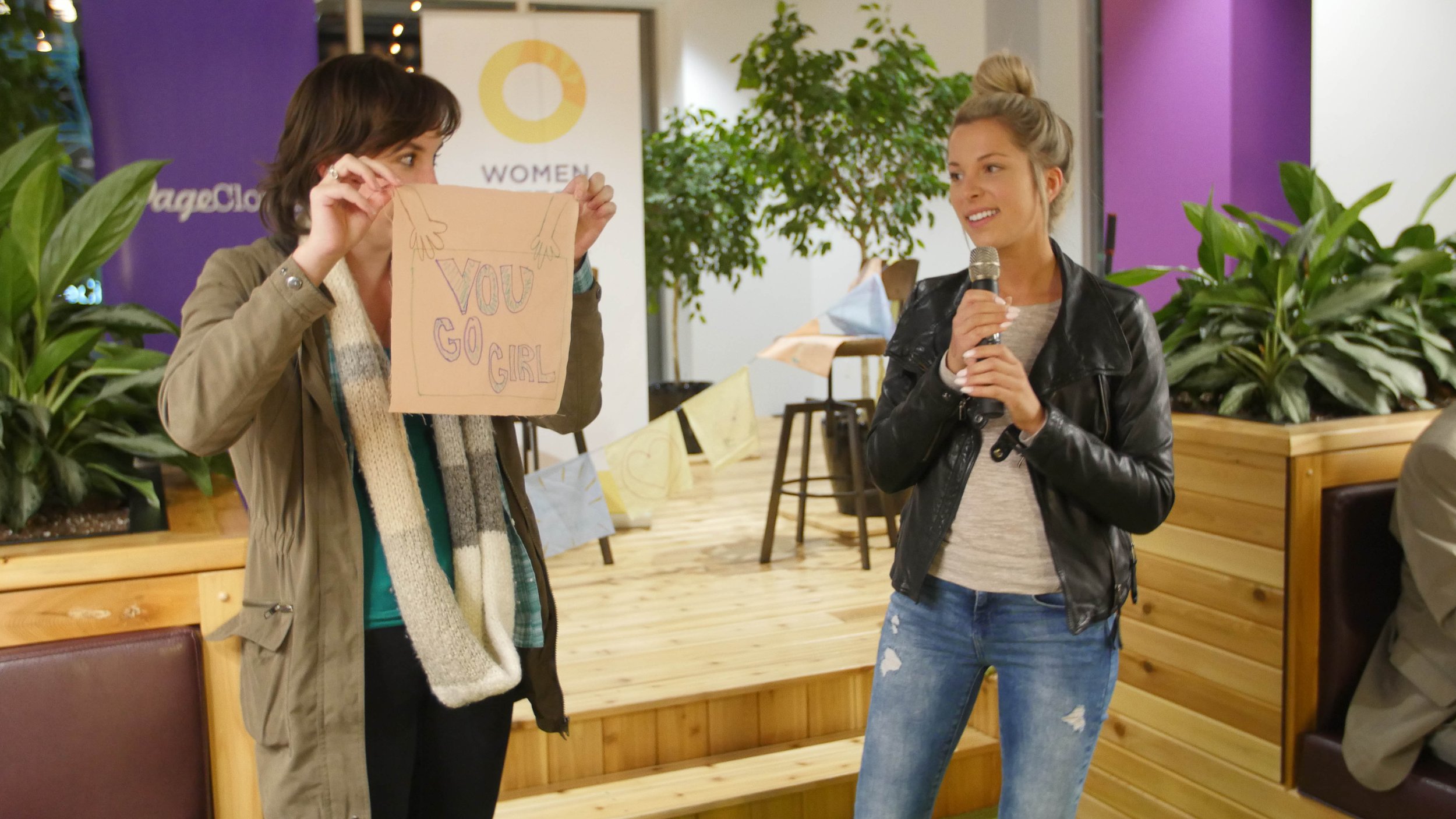  Two women are standing in front of the audience, one has a mic and the other is holding a piece of paper that says “You Go Girl.”  