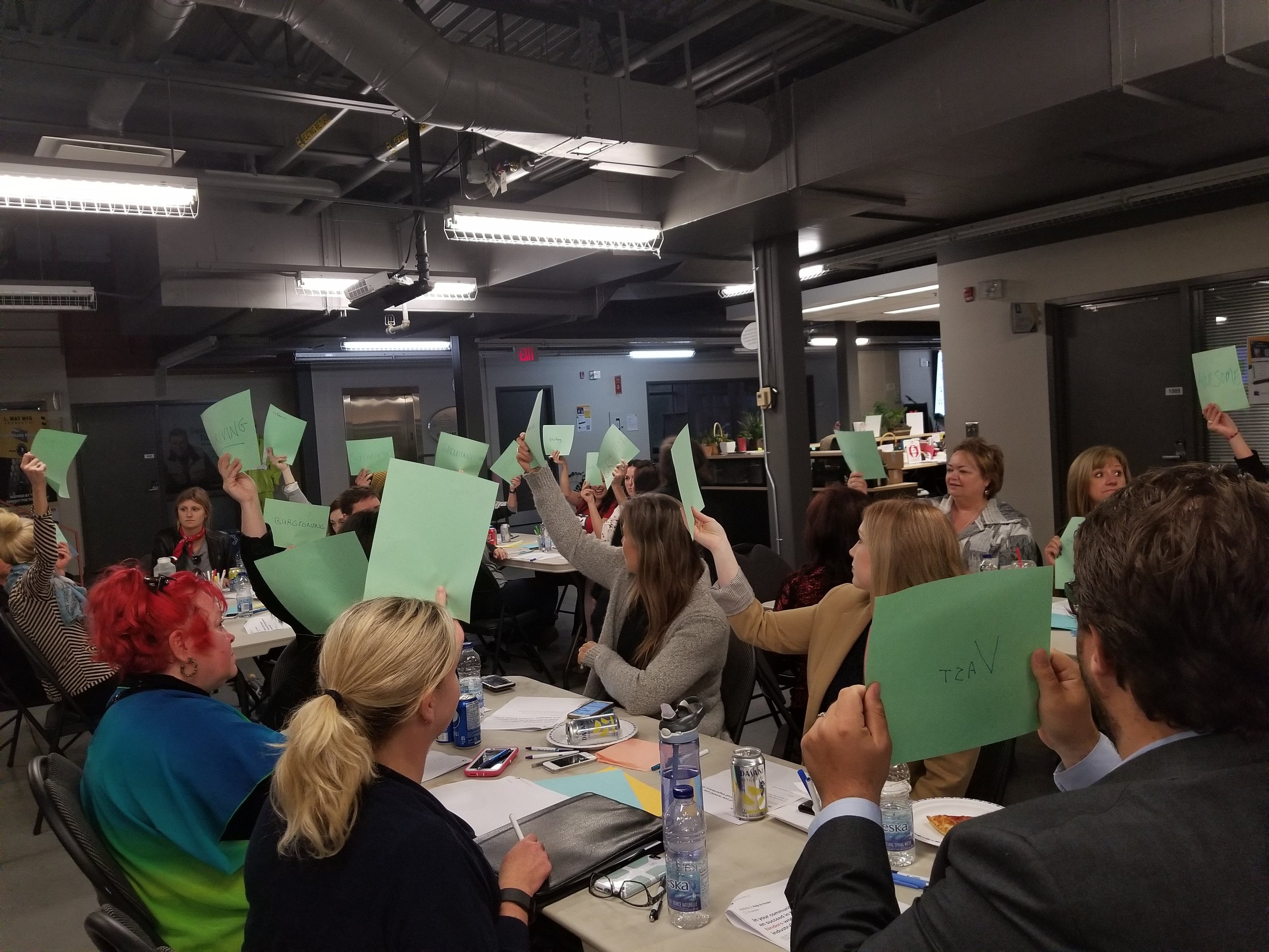  Image is of a group of people sitting down at the Community Conversation in Sudbury, Ontario. The audience is holding up green sheets of paper with ideas written on them.  