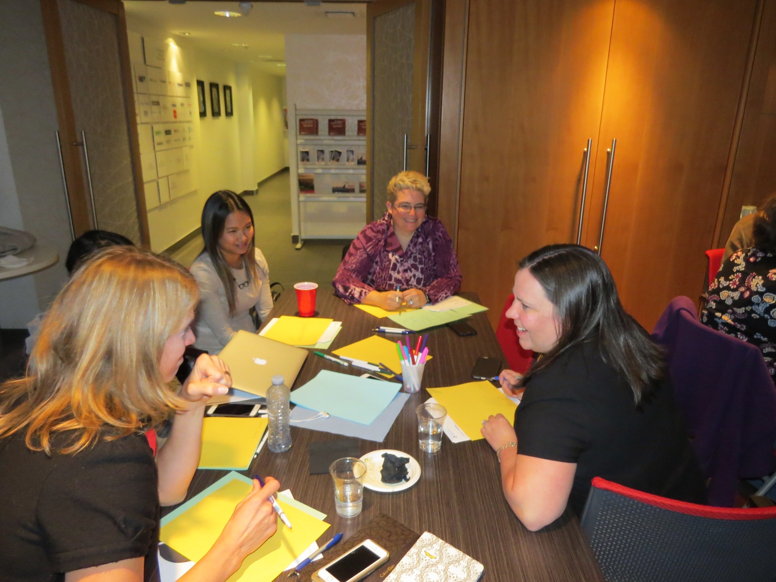  Image is of several woman working at a table together, writing things down on yellow pieces of paper and chatting at the Community Conversation in Calgary. 