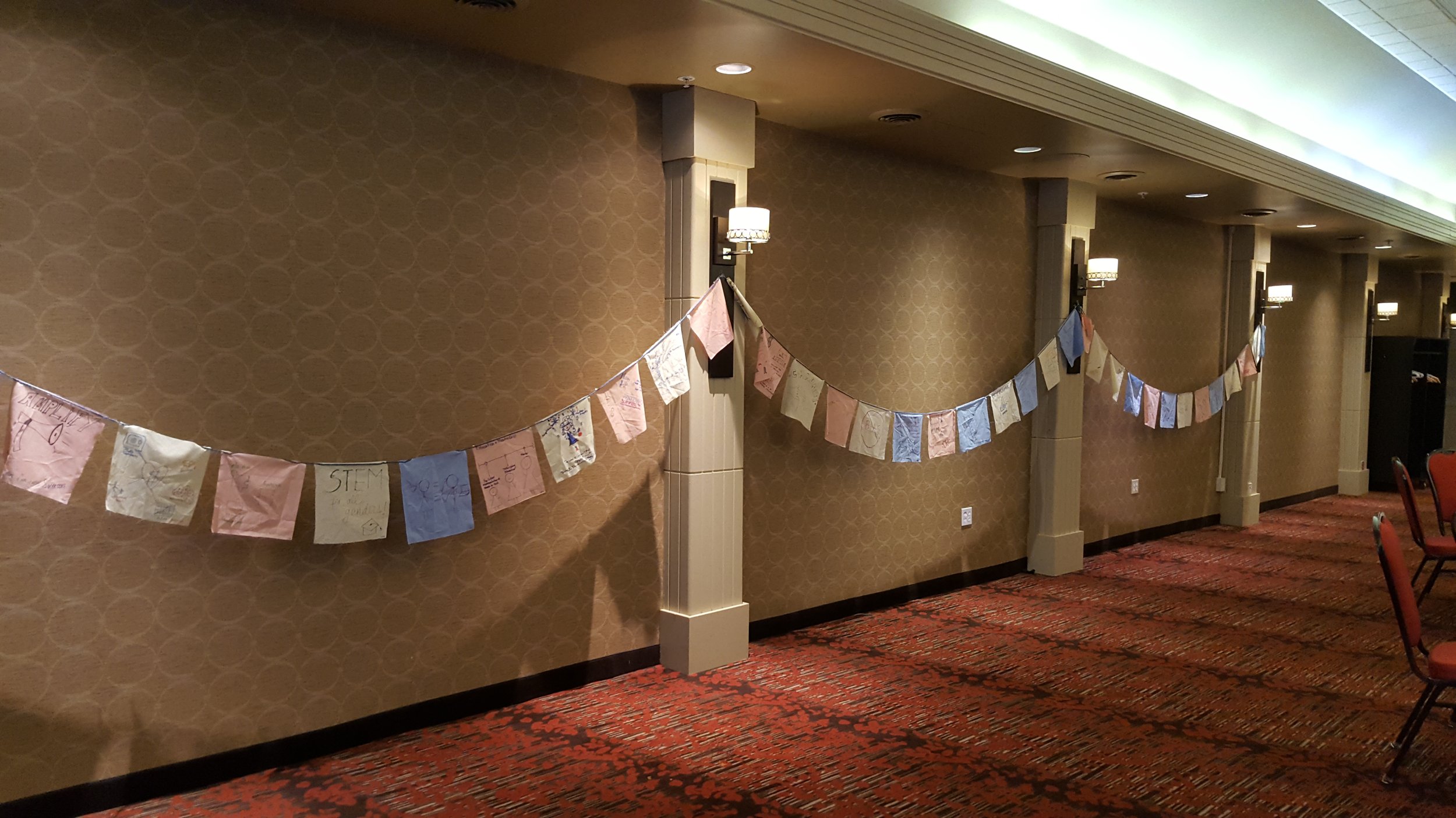  Image is of sheets of paper hanging by a string going from wall to wall in a “u” shape. The pieces of paper of the ideas from the Community Conversation written on them. 