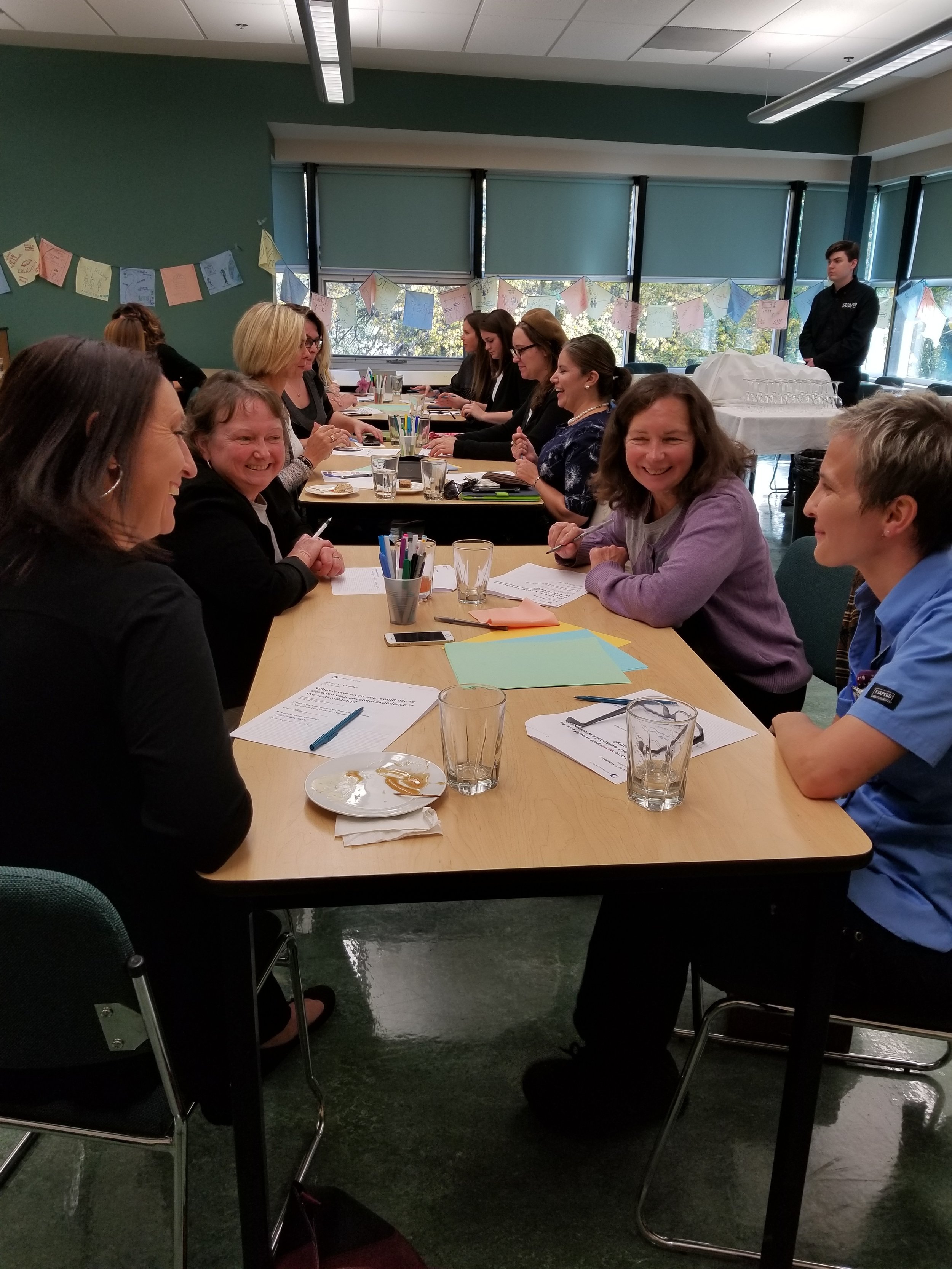  Image is of people gathered at a table for the Community Conversation in Brandon. They have paper and pens on the table along with plates of food and glasses of water. The women at the table seem to be smiling and having a conversation with one anot