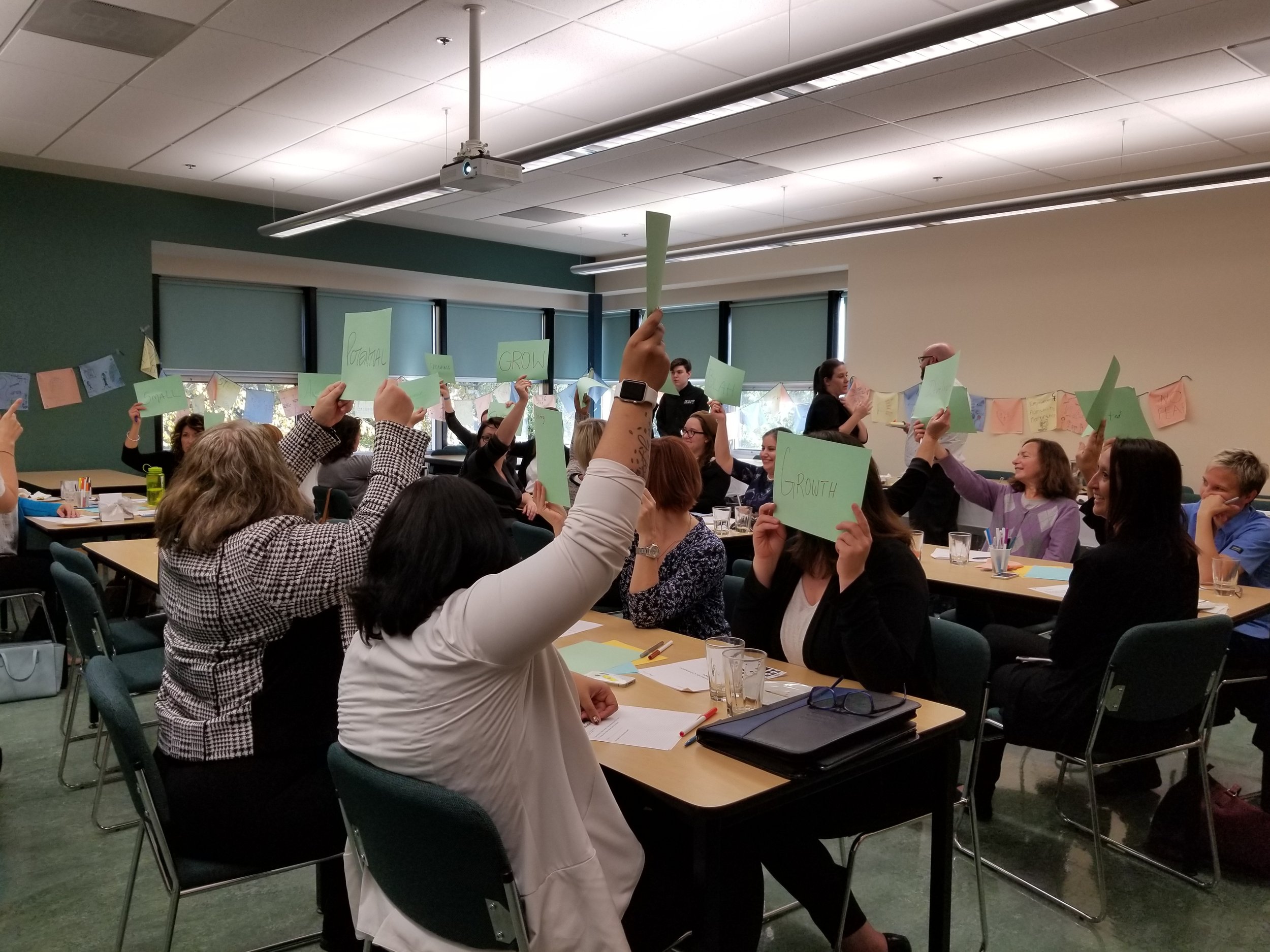  Image is of the Community Conversation in Brandon. People in the room are sitting at tables and everyone is holding up a green sheet of paper with something written on it. 