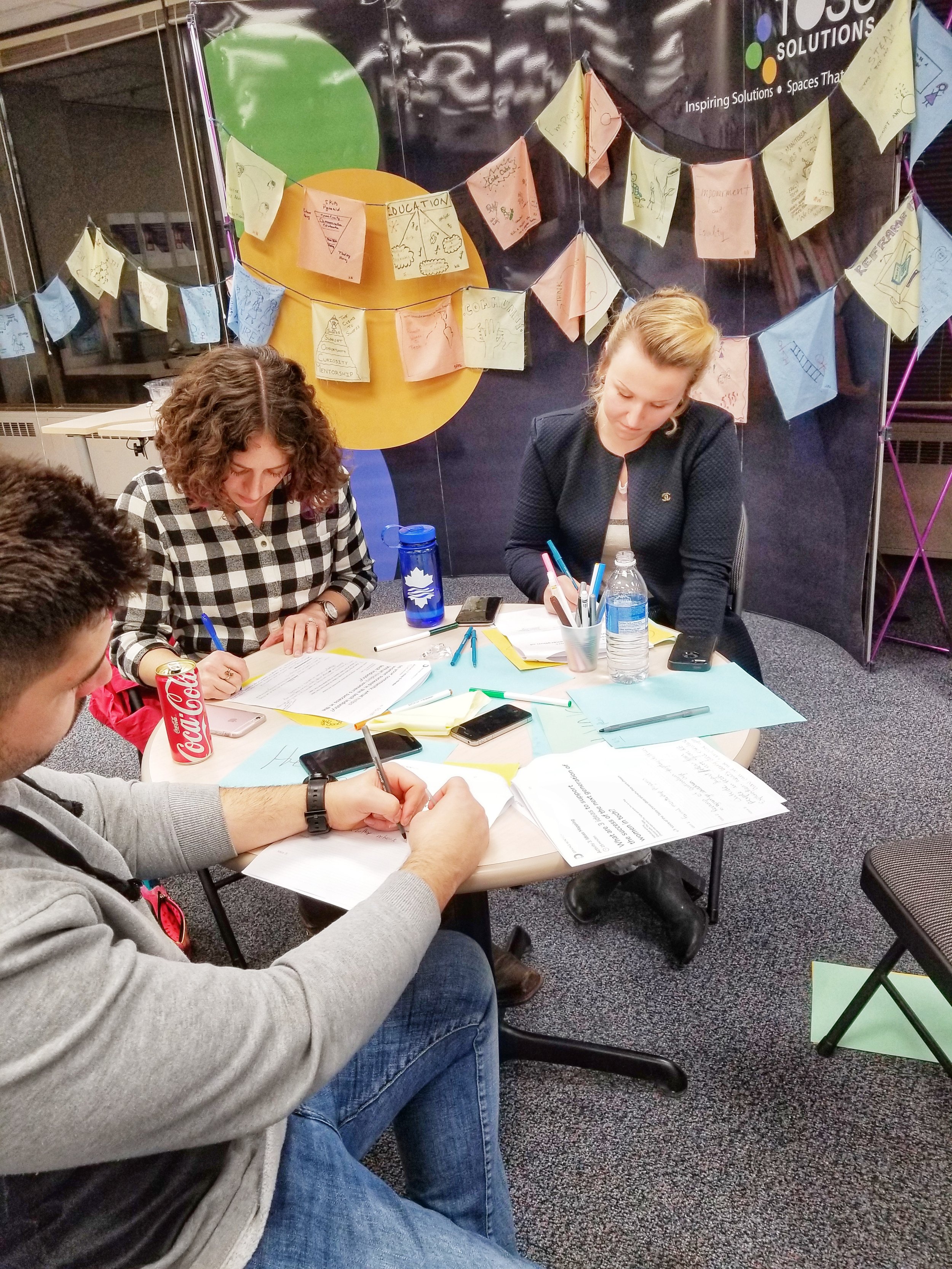  Image is of a group of three people at the St. John’s Community Conversation sitting at a table all writing something down on the worksheets they were provided. 