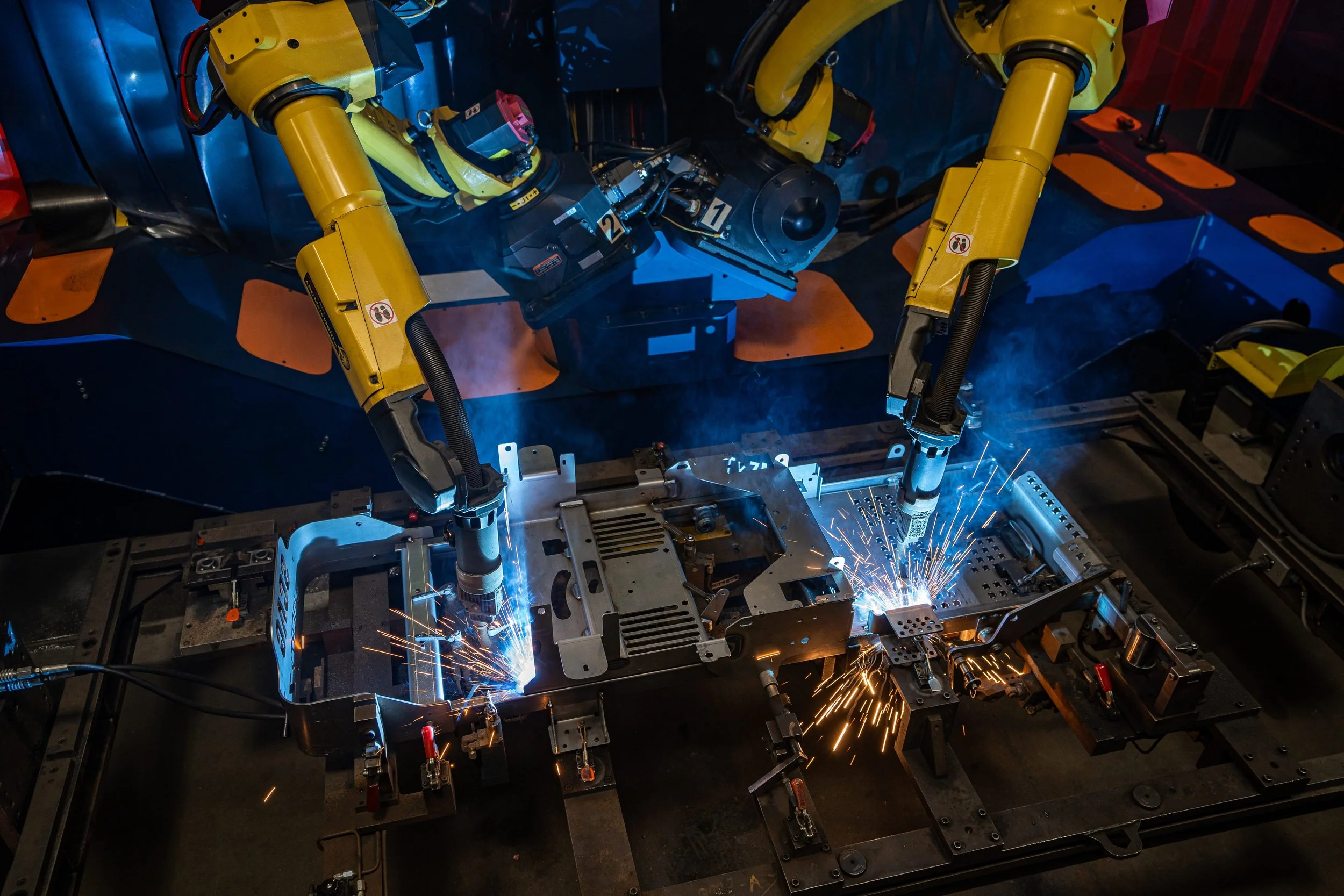 Two yellow robotic arms working on an assembly line. Sparks are flying off from the ends of the robotic arms.