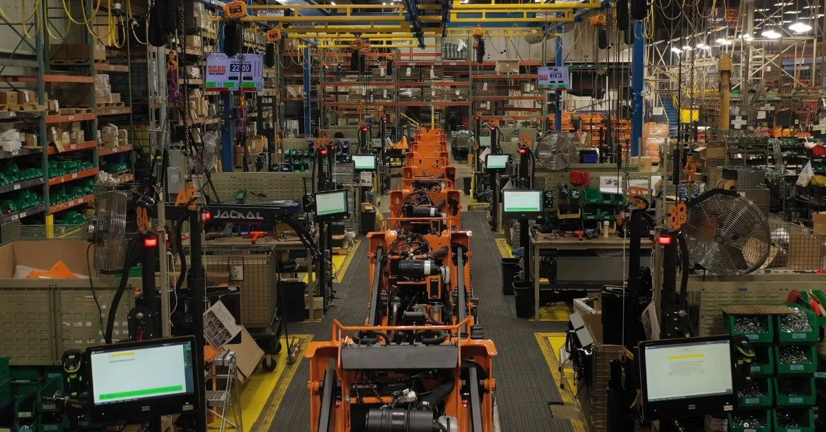 The interior of a massive assembly line warehouse. There are mechanical pieces scattered all around the warehouse.