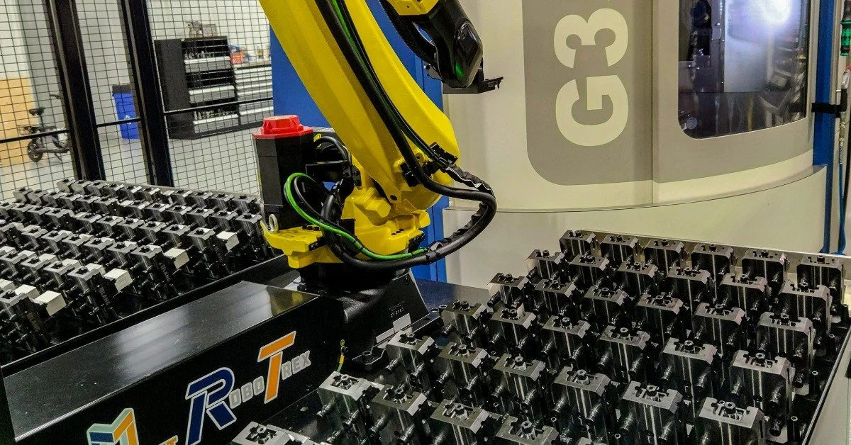 A yellow robotic arm reaching between rows of black metal parts beside an enclosed industrial machine.