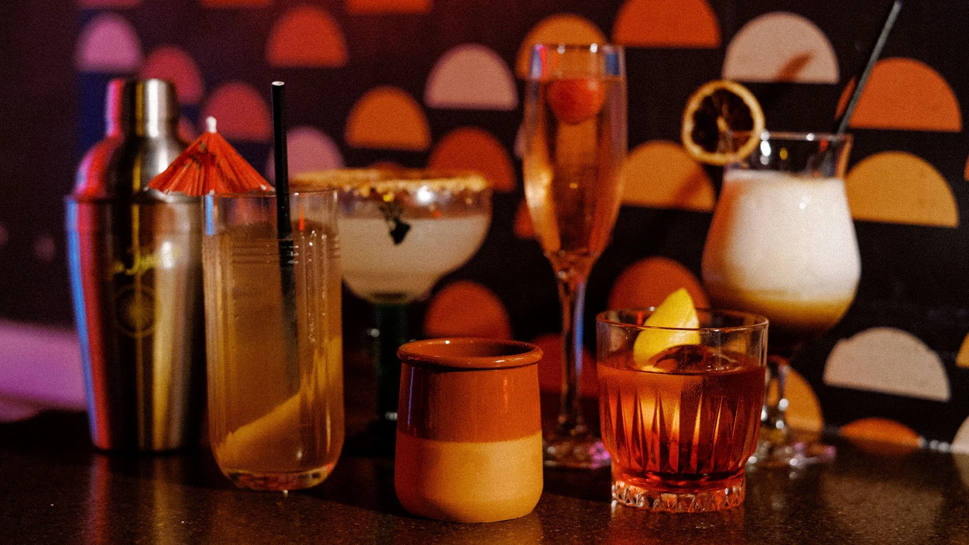 Various cocktails and glasses with garnishes on a bar counter with a colorful, semi-circular patterned background.