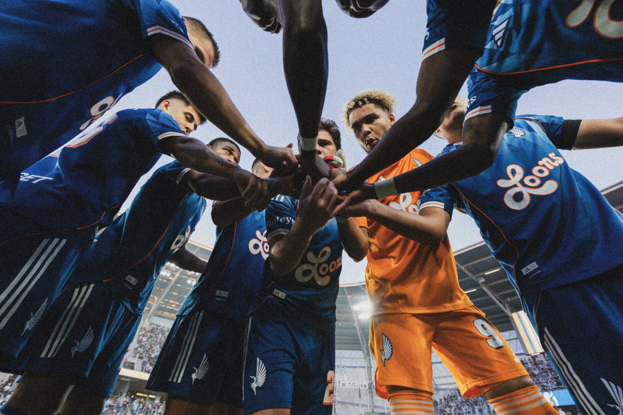 Soccer players in blue and orange uniforms gathering in a huddle on the field.