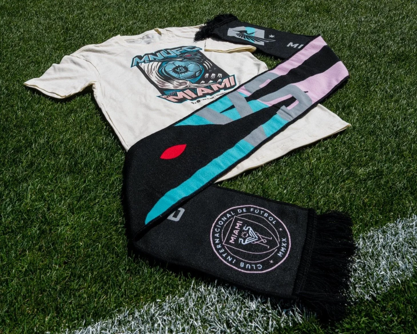 A white t-shirt and a black scarf with Minnesota United FC and Inter Miami football team logos lying on a grassy field.