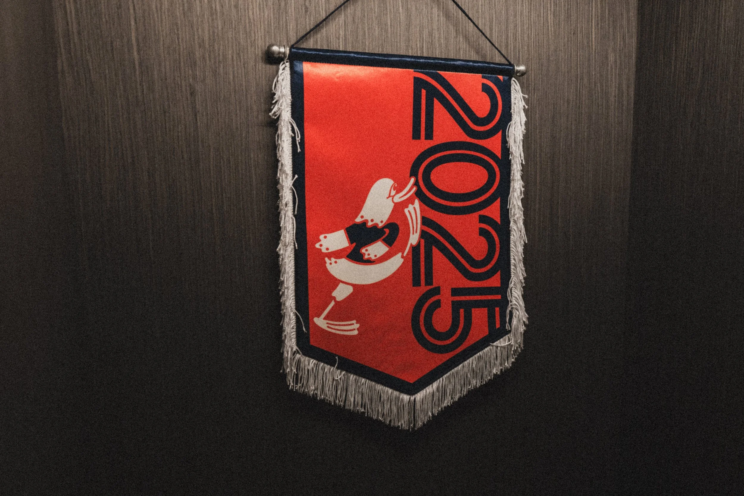 A decorative banner with an orange and navy blue design, featuring a loon and the year '2025' on fabric with fringe trim, hanging on a wooden wall.
