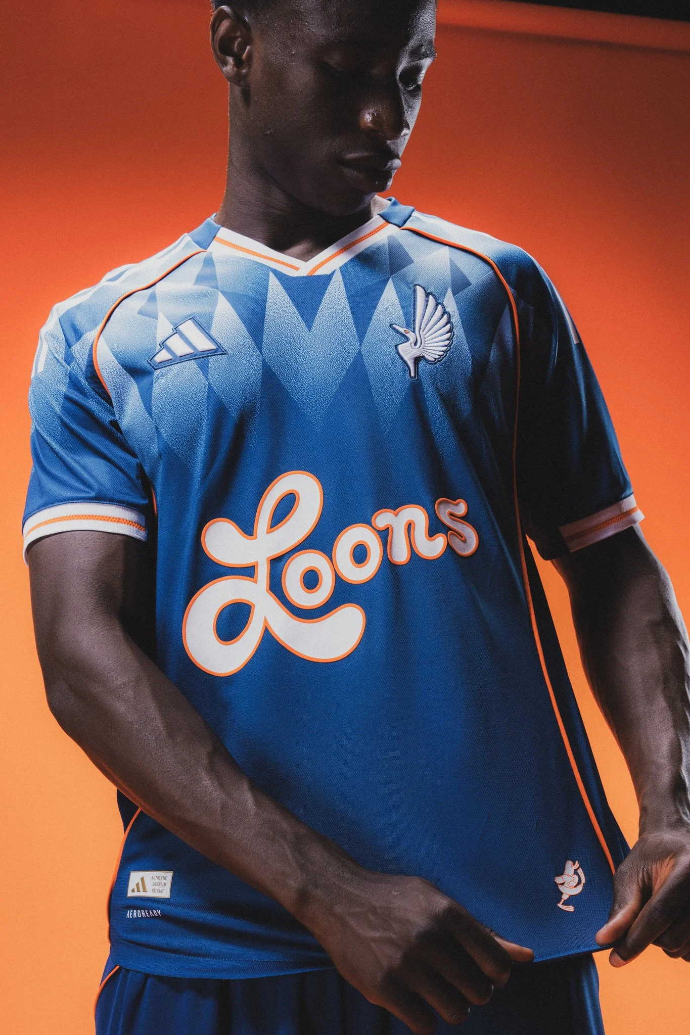 A professional soccer player wearing a blue sports jersey with orange accents, featuring a logo of a loon on the chest and a patch on the bottom left. The background is orange, and the person's arms are crossed.