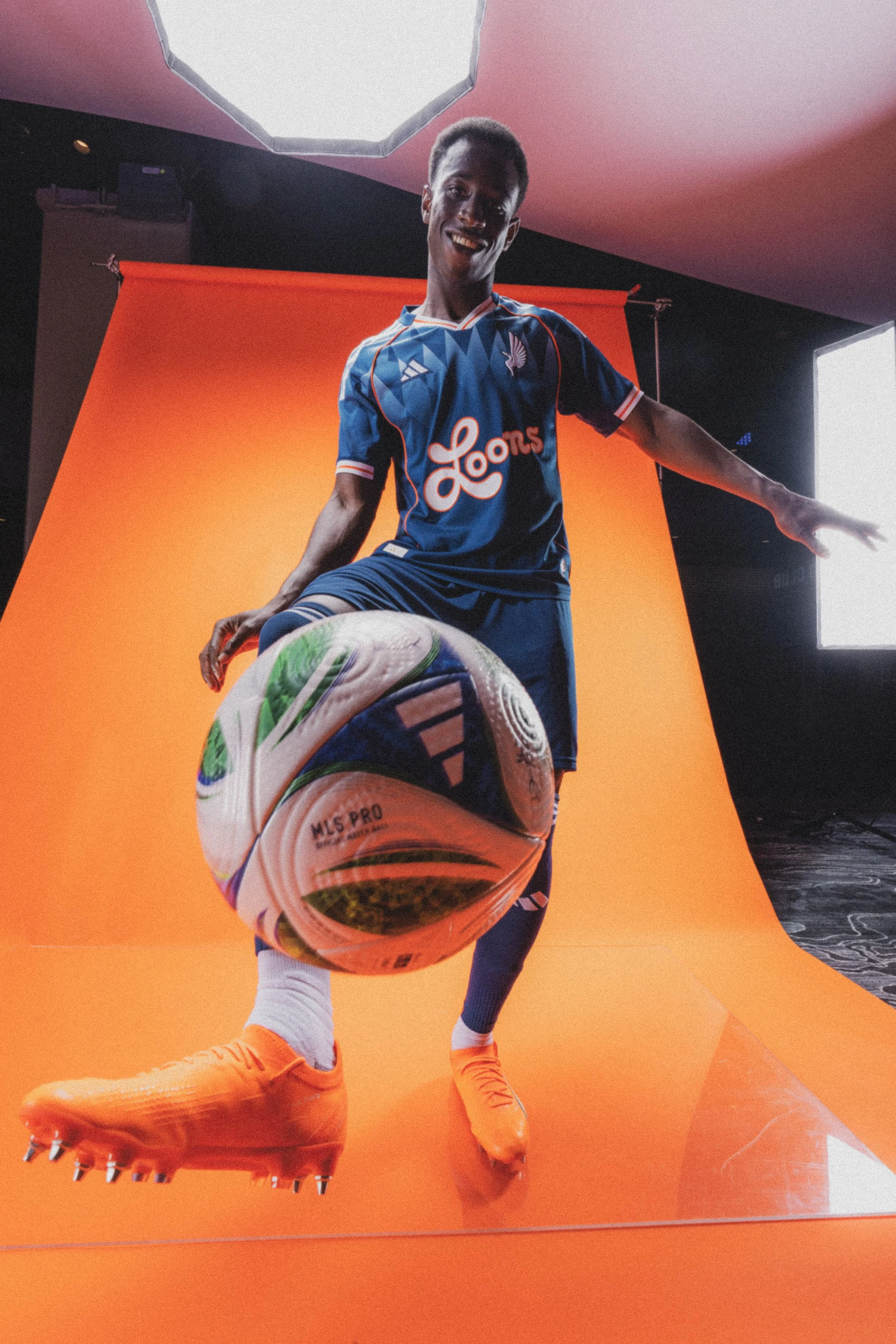 A male athlete wearing a blue soccer jersey and orange cleats kicks a soccer ball on an orange background in a studio setting.