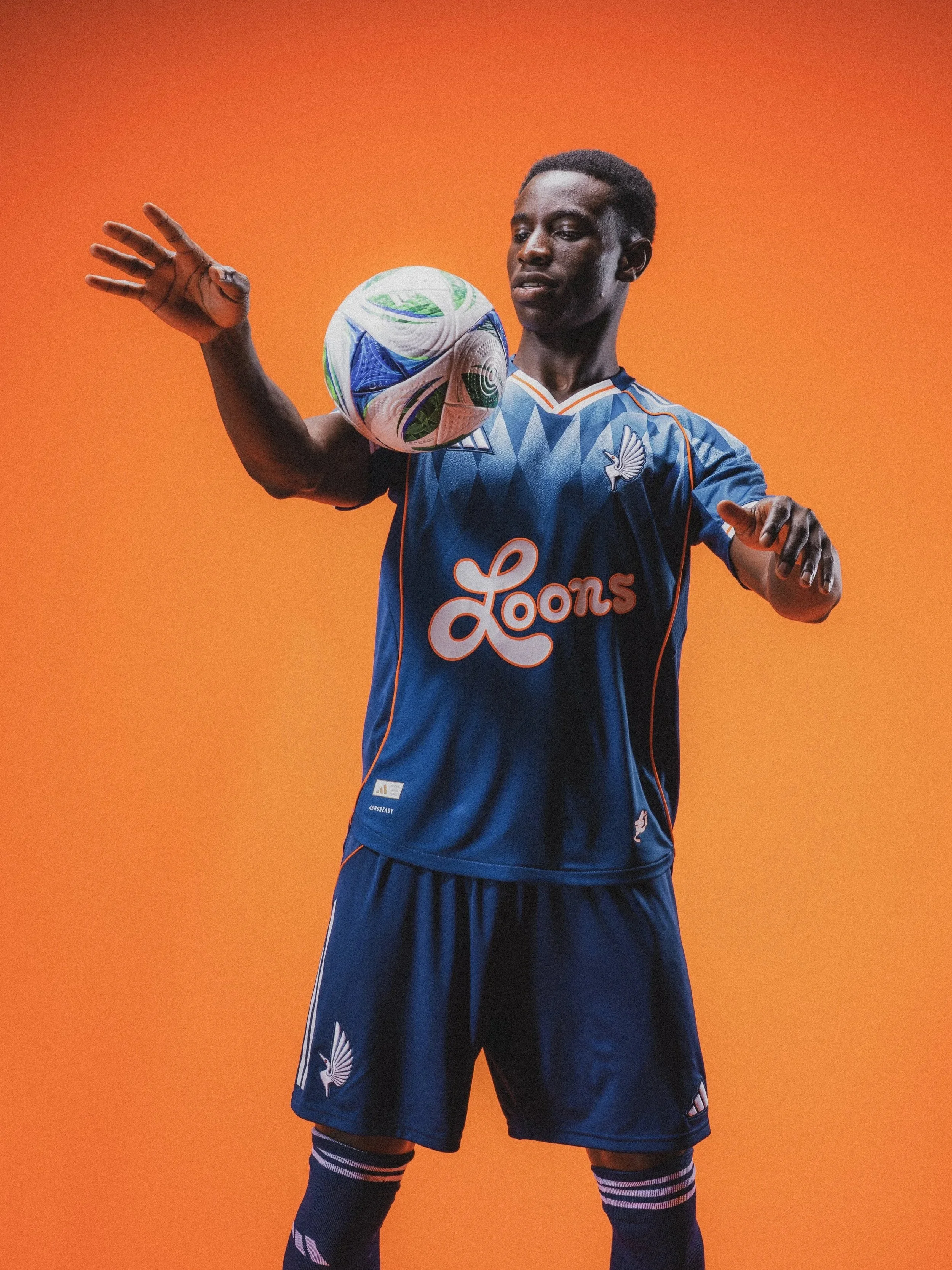 A male soccer player in a blue jersey with white and orange details, holding or balancing a multicolored soccer ball on his shoulder, standing against an orange background.