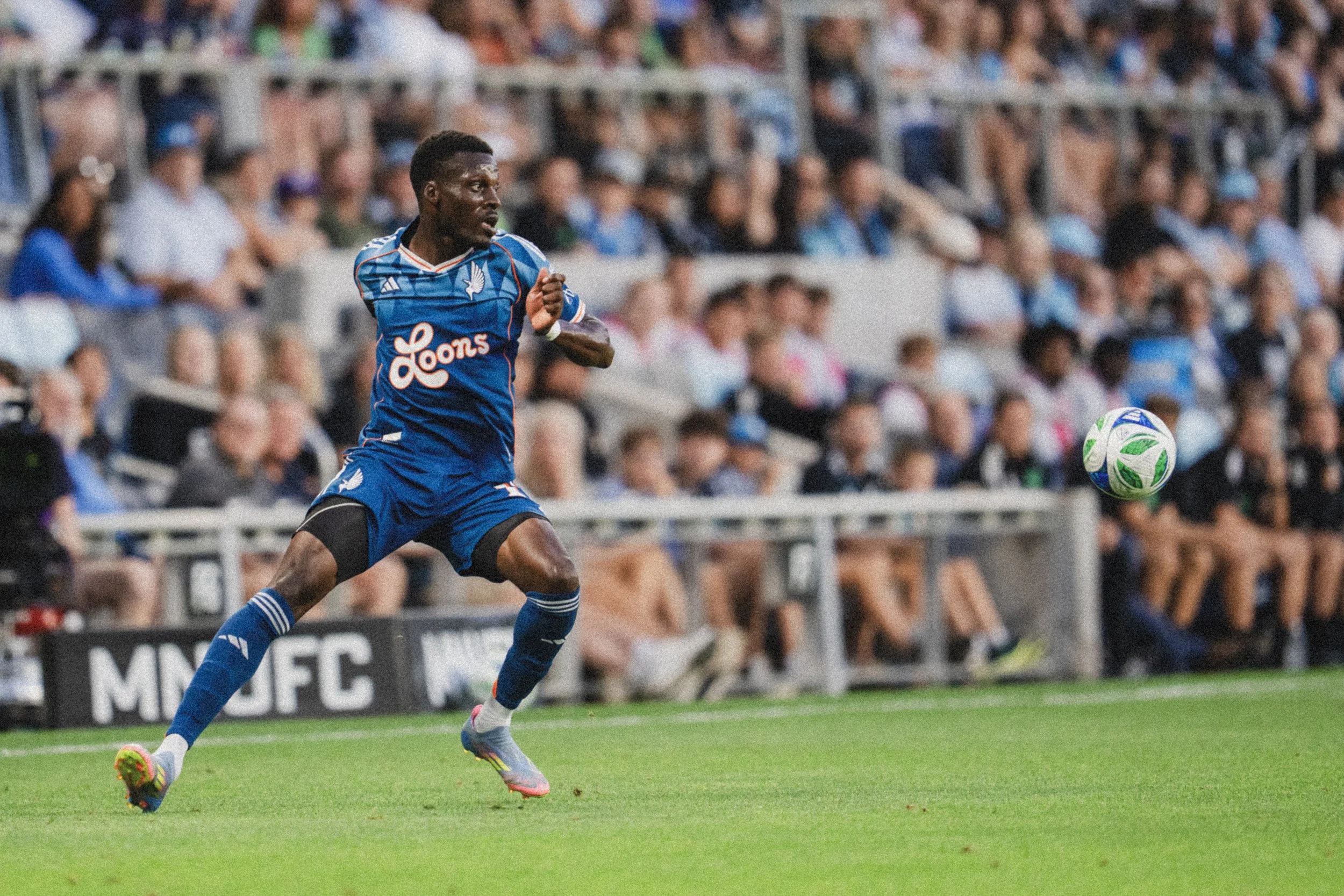 A soccer player in a blue uniform,  running on the field with a white soccer ball in front of him during a match. The crowd is visible in the background.