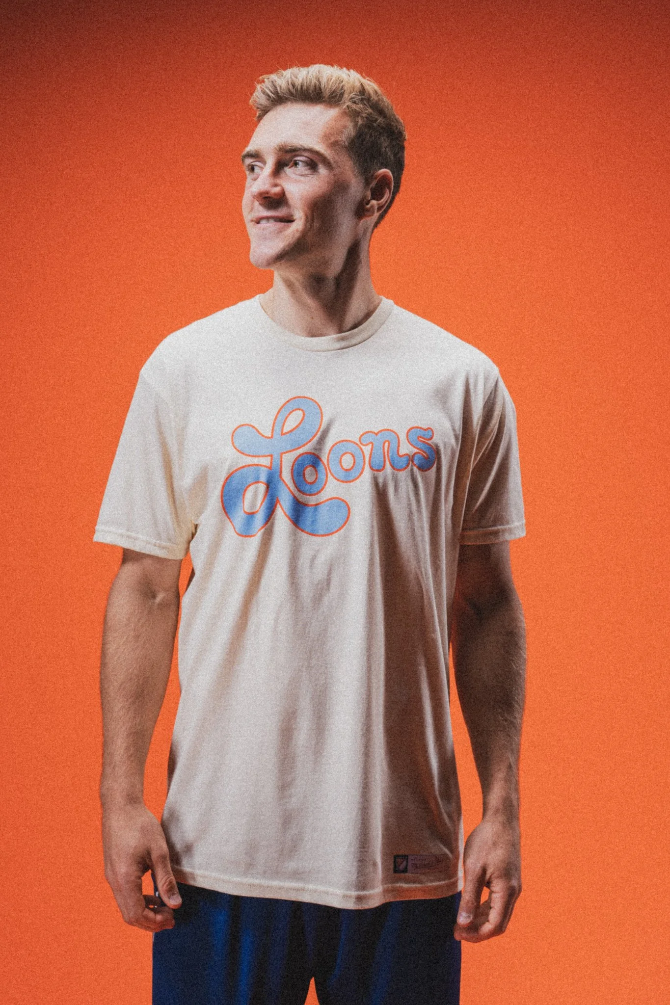 A young man with short blonde hair wearing a beige T-shirt that says "Loons" in colorful cursive lettering, standing against an orange background.