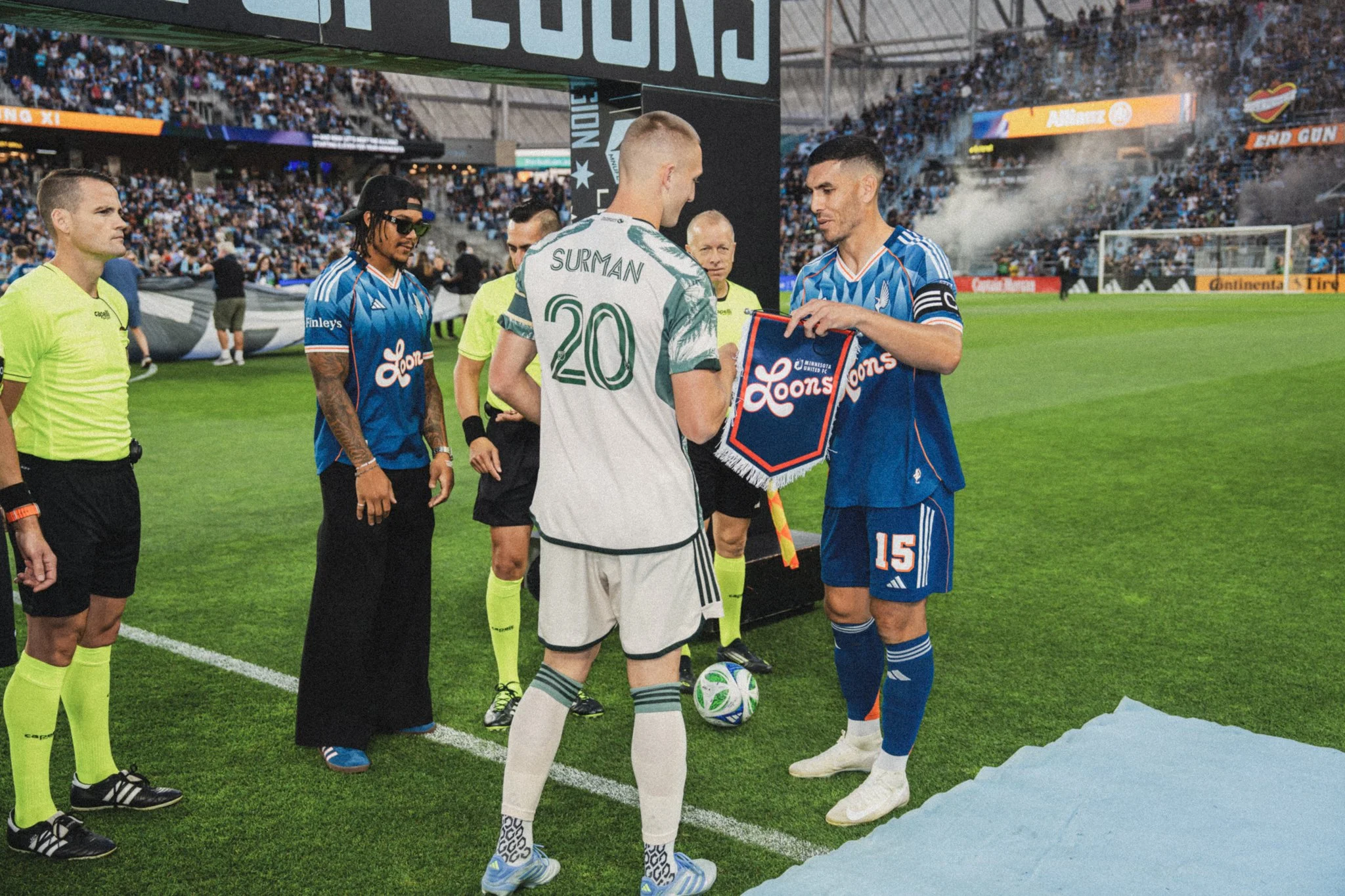 Soccer players and officials standing on the field before a match, with a referee and two team captains shaking hands, crowd in the stadium stands.
