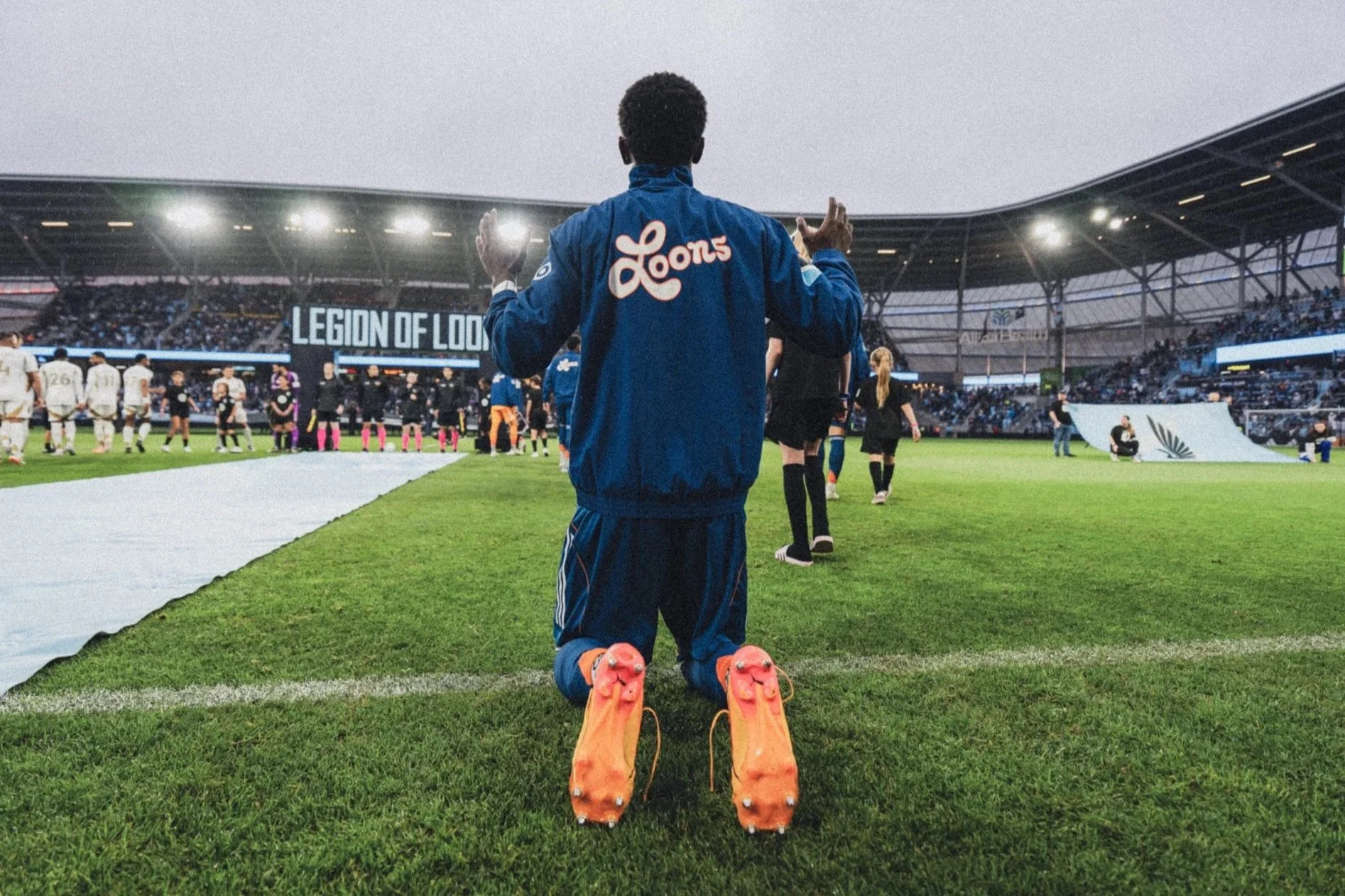 Soccer player kneeling on the field with orange cleats, watching players and referees before a match at a stadium.