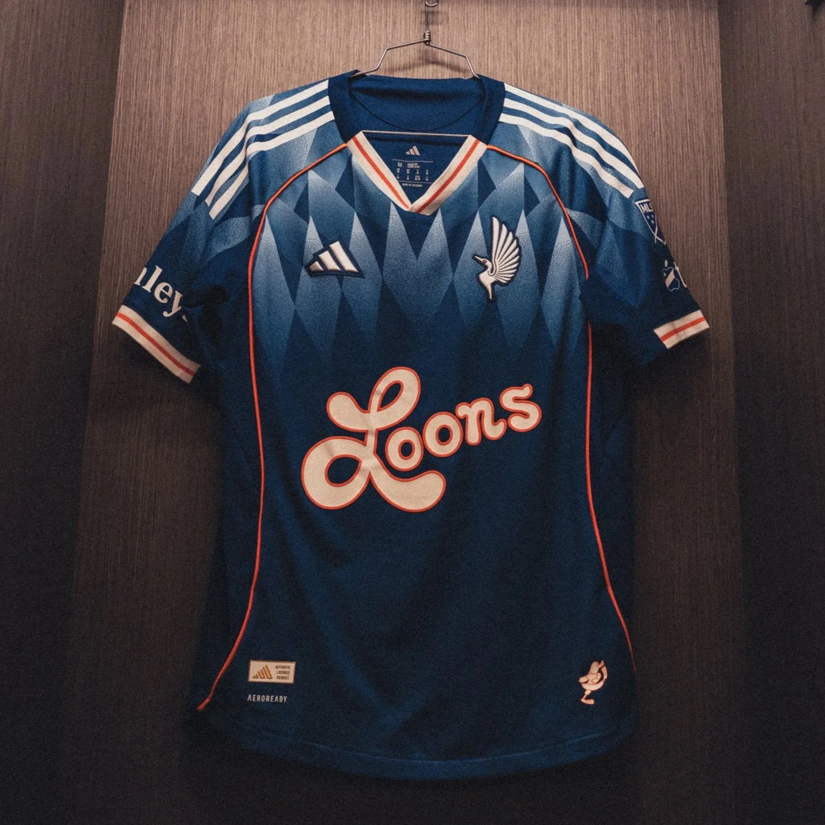 Blue soccer jersey hanging on a hanger in a locker. The jersey features a geometric pattern, the Minnesota United FC logo, and large script that says 'Loons'.