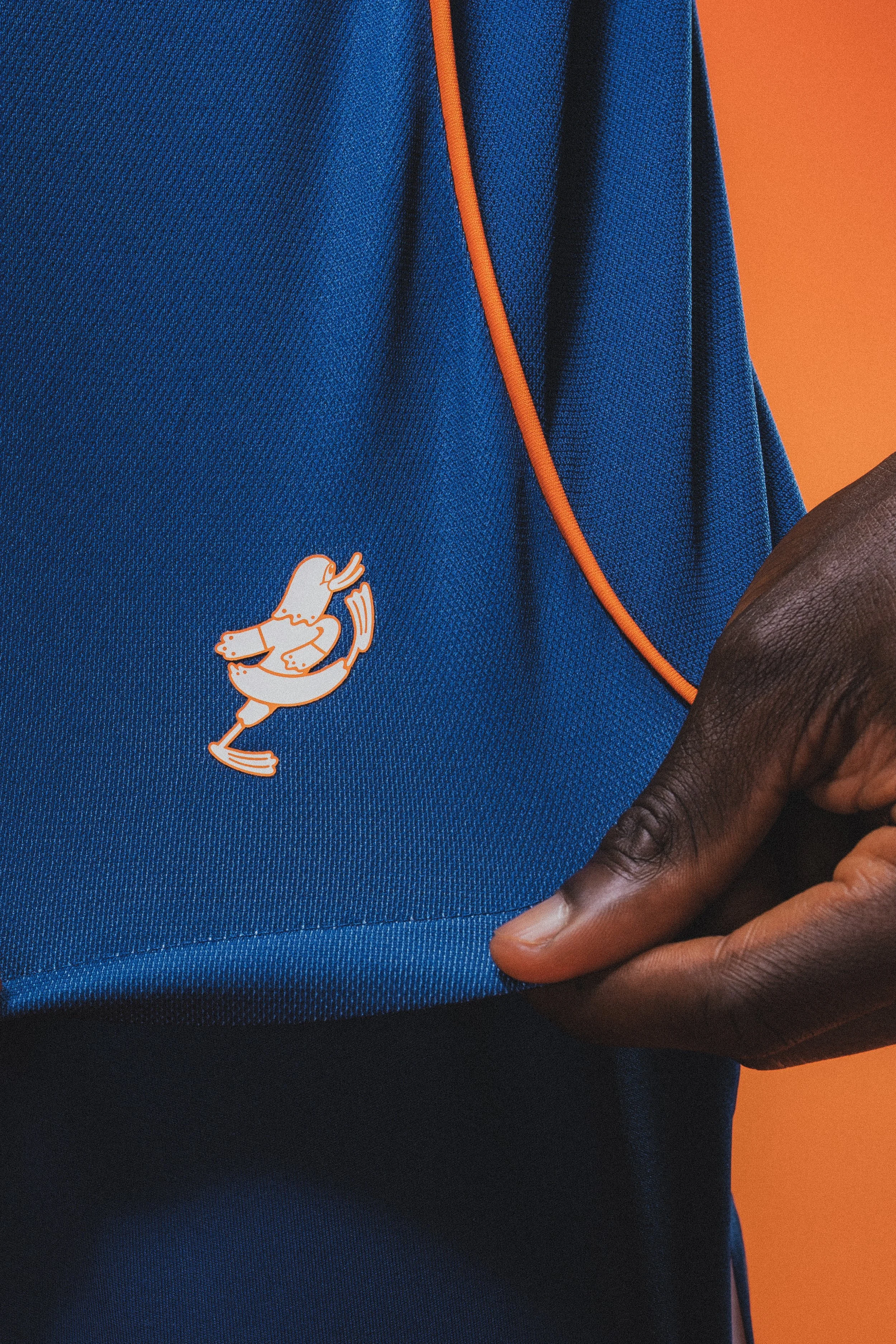 Close-up of a blue soccer jersey with orange piping and a white loon mascot, held in someone's hand against an orange background.