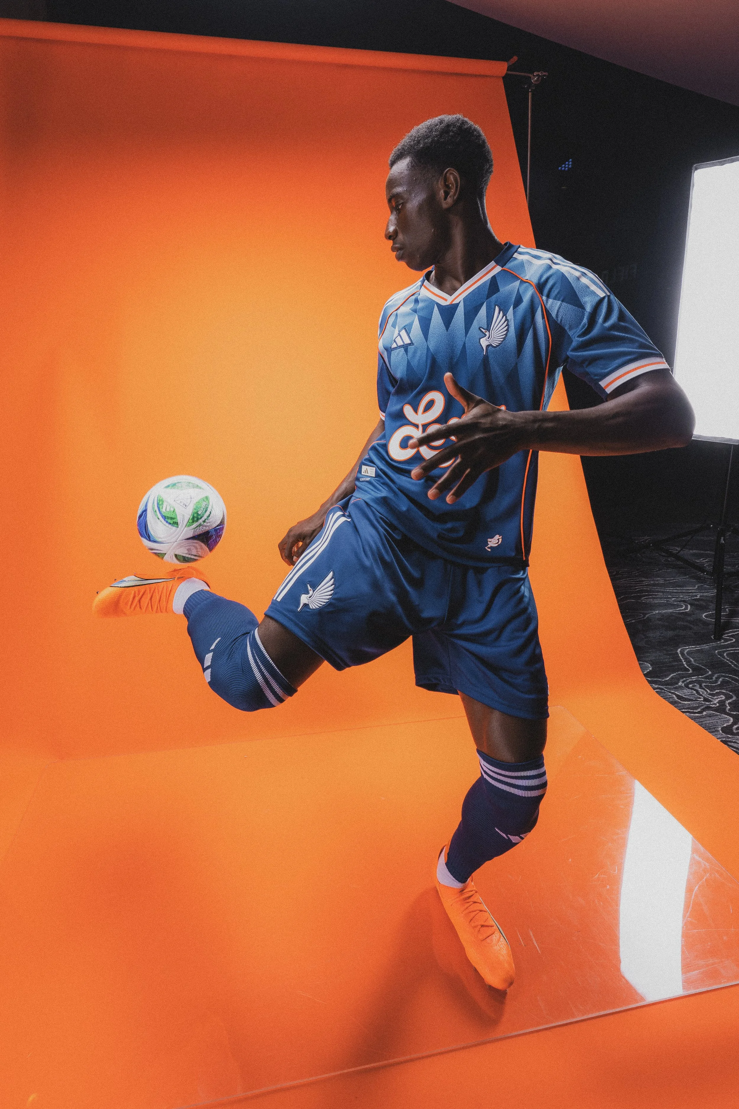 A male soccer player in a blue jersey, orange cleats, and navy socks, balancing a soccer ball on his foot against an orange backdrop in a photo studio.
