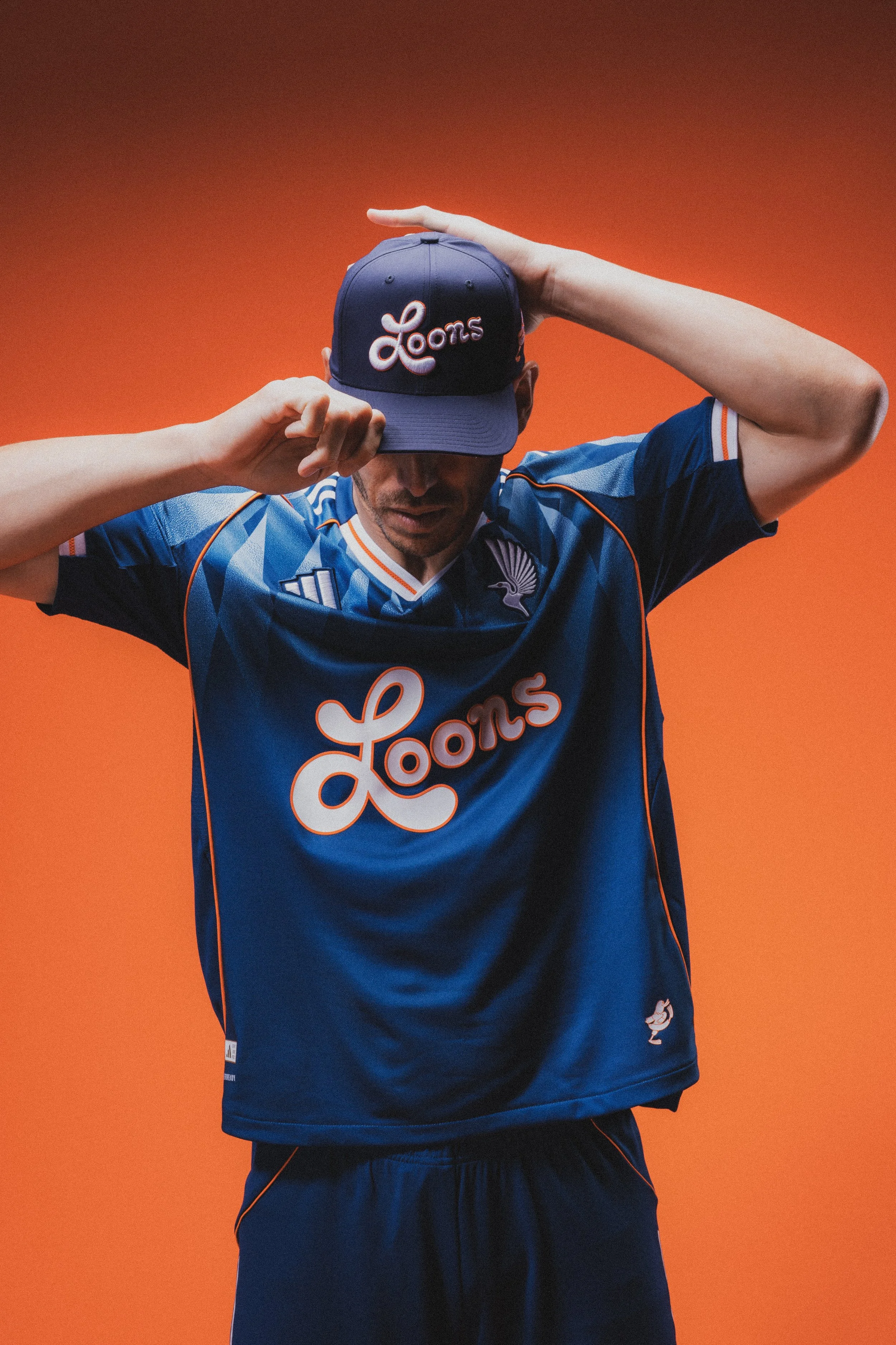 A man wearing a blue sports jersey and matching cap with the logo 'Loons' adjusting his cap, against an orange background.