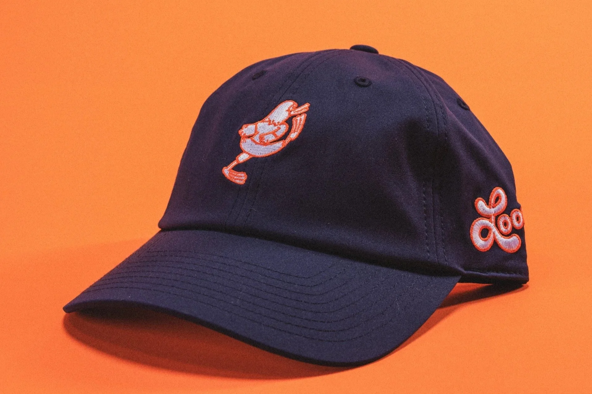 A dark blue baseball cap with orange and white embroidered loon mascot on the front and side, placed on an orange background.