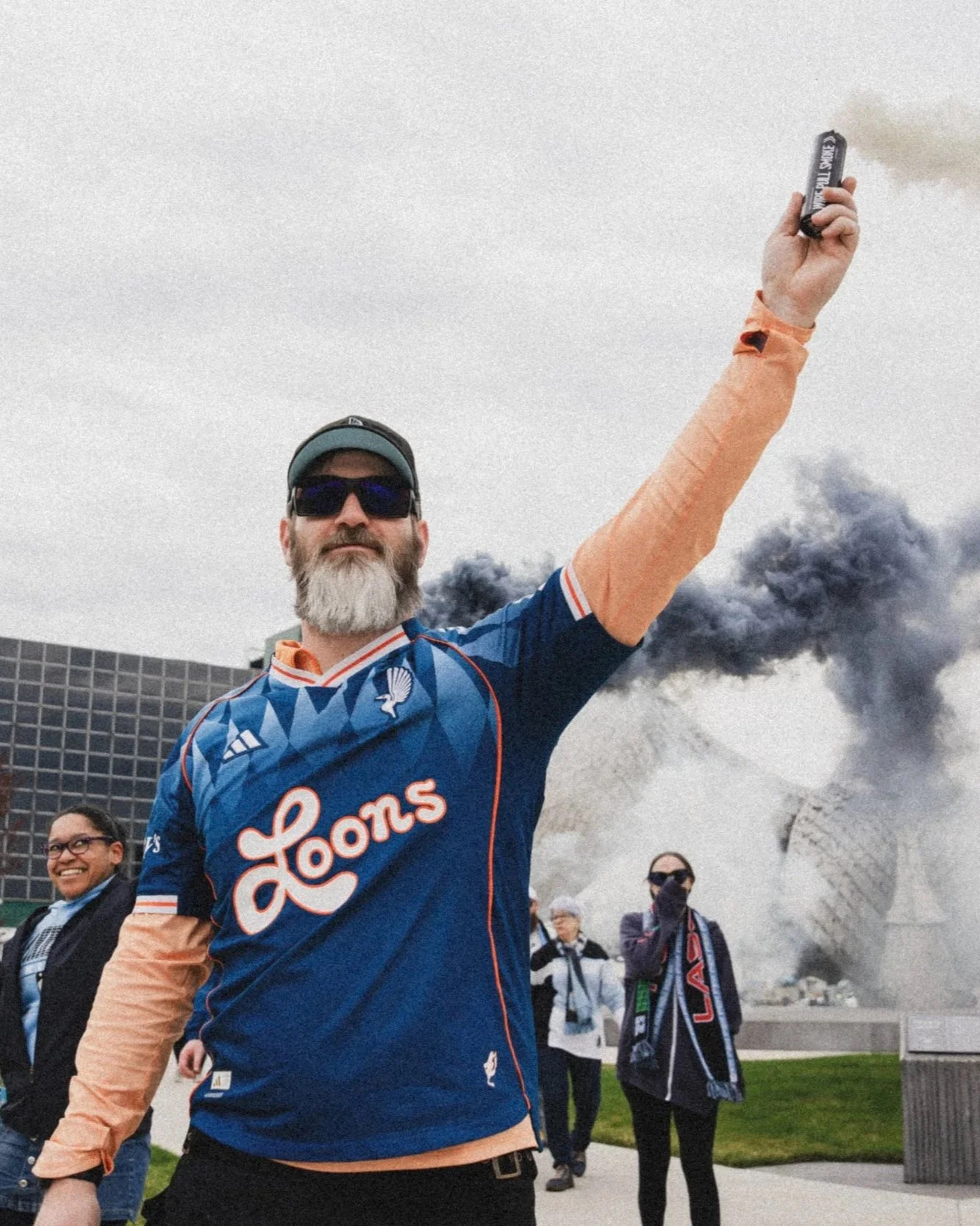 A man wearing sunglasses, a baseball cap, and a blue sports jersey is holding a smoke grenade that is emitting black smoke. Several people are in the background, and there is a large building with smoke or steam rising behind them.