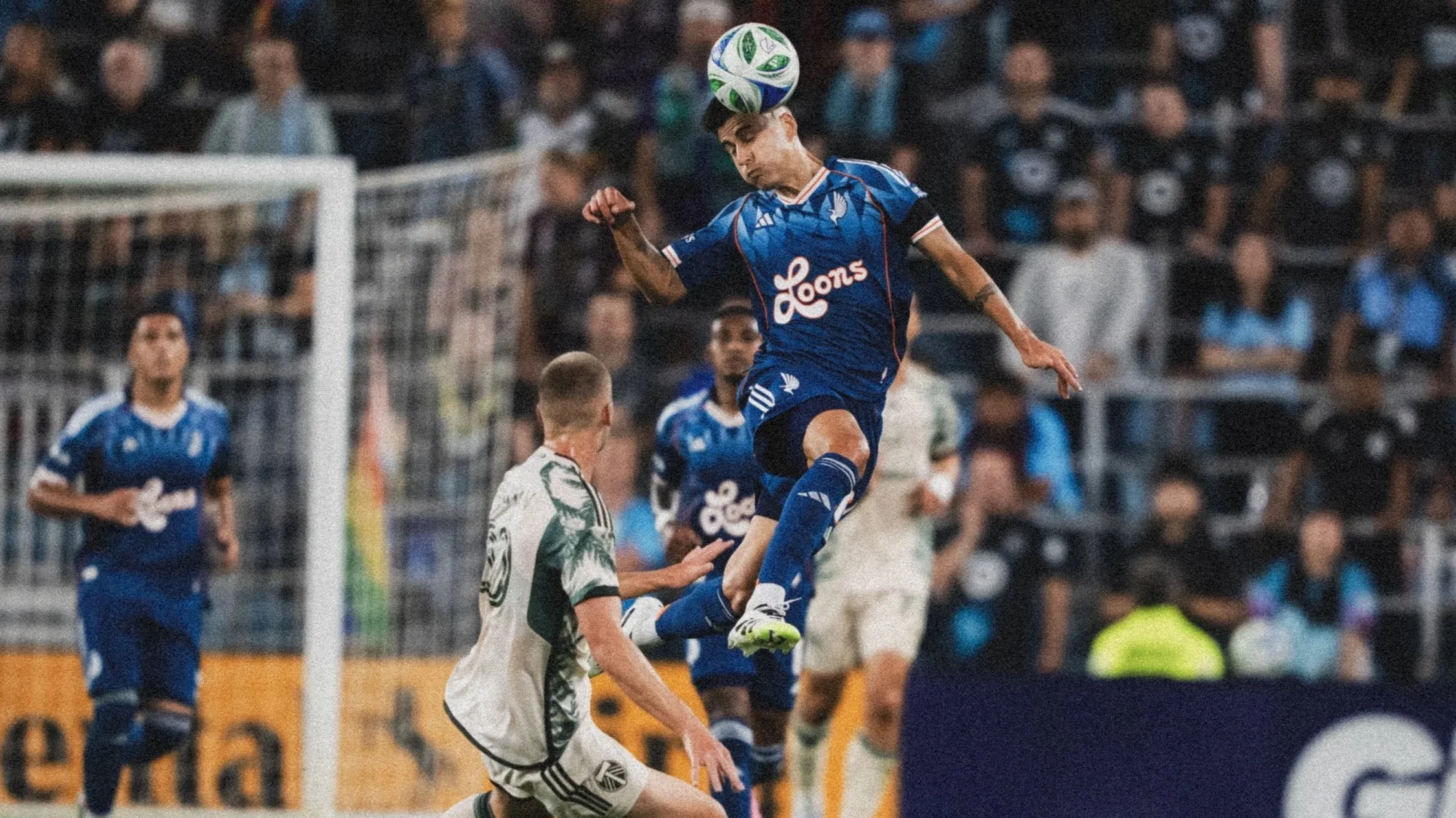 A soccer player in a blue uniform jumping to head the ball during a game, with a crowd of spectators in the background.