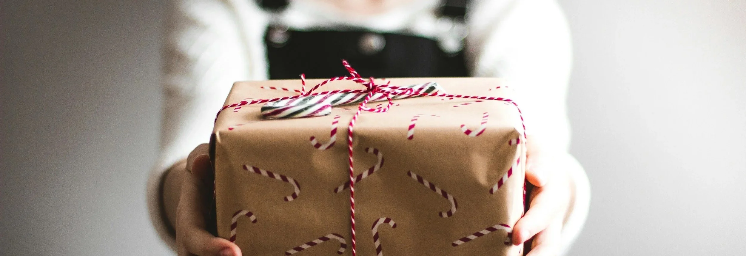 Person holding a wrapped gift box decorated with candy cane patterned paper and tied with red and white twine. Gift cards are perfect for every occasion.