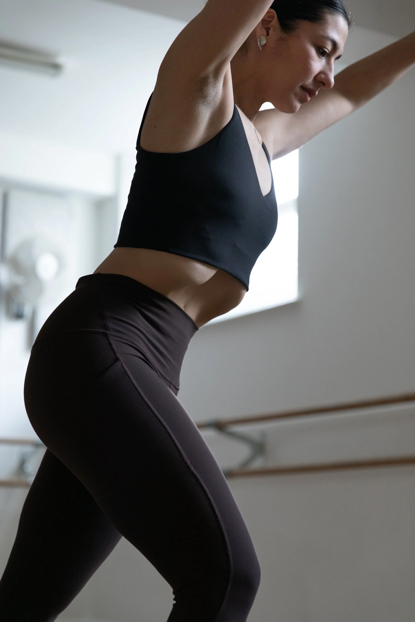 Woman performing a Hypopressive pose on a black mat in a room with light-colored wooden flooring and plain white walls.