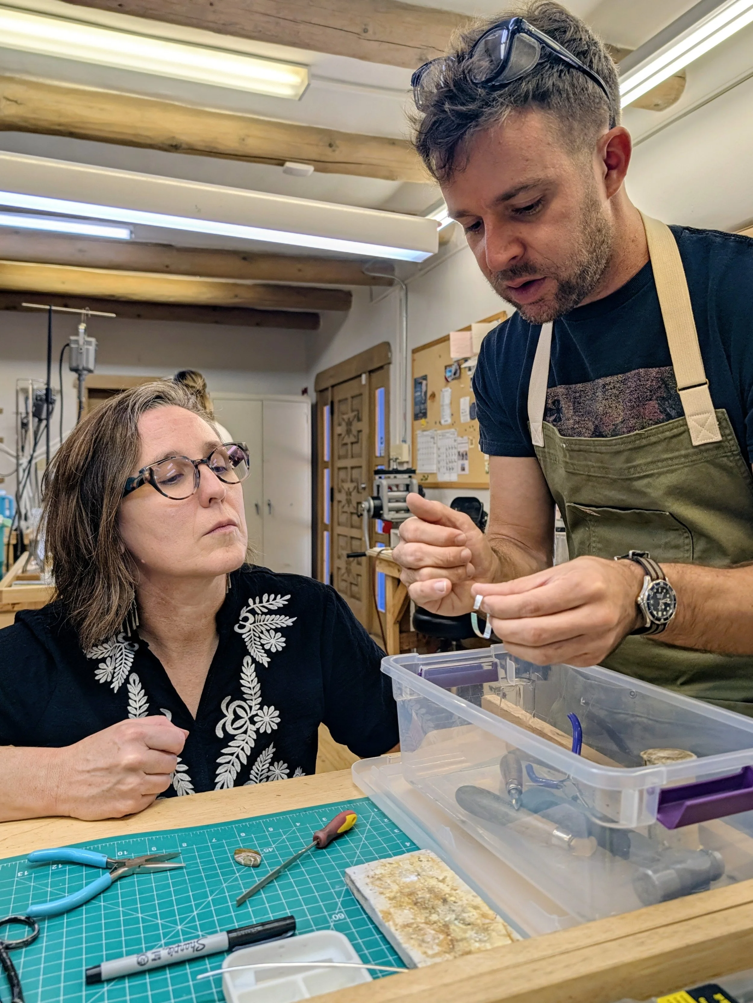 Instructor demonstrating a jewelry-making technique to a student at the workbench