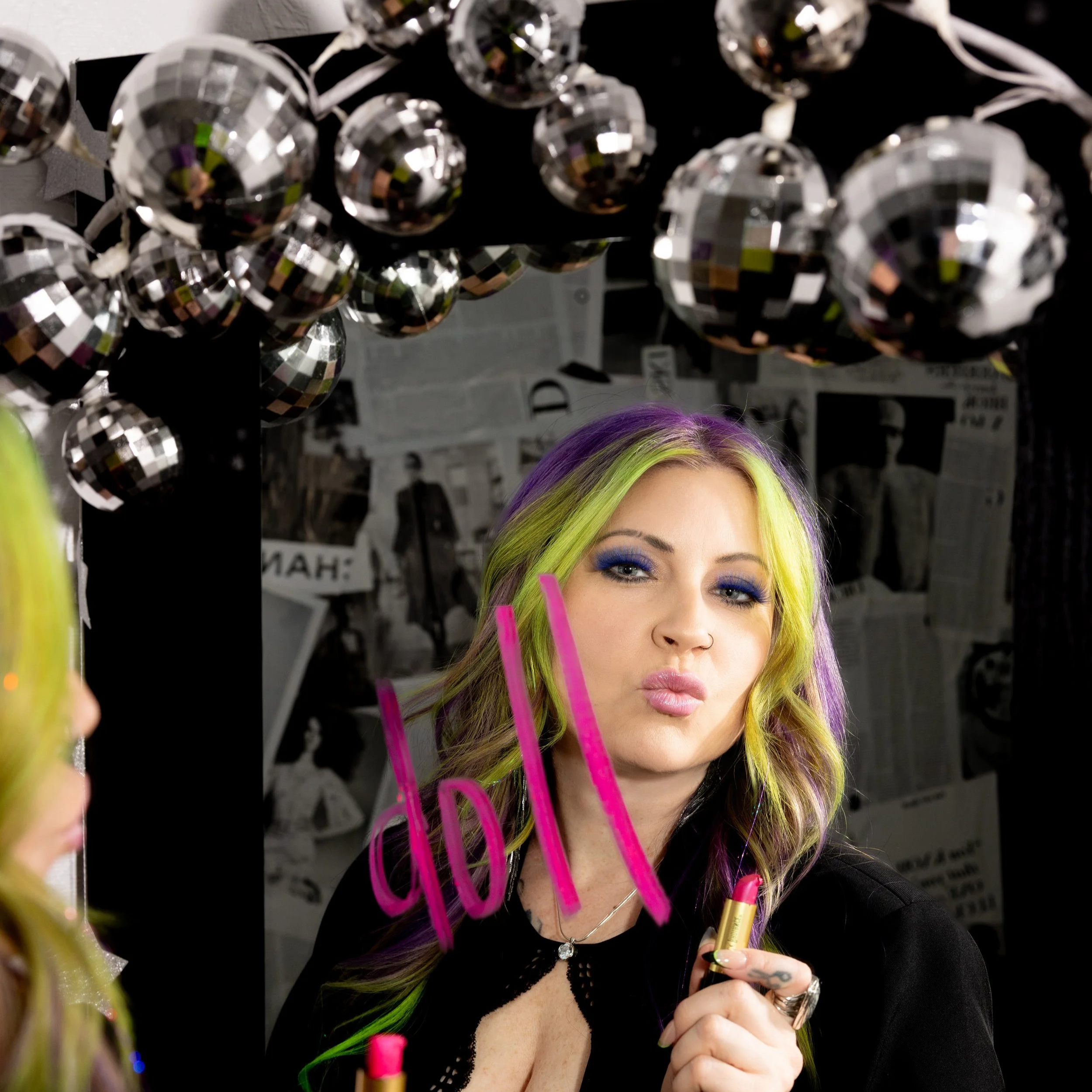 a hairstylist surrounded by disco balls blowing a kiss, the word "doll" written in pink lipstick on a mirror